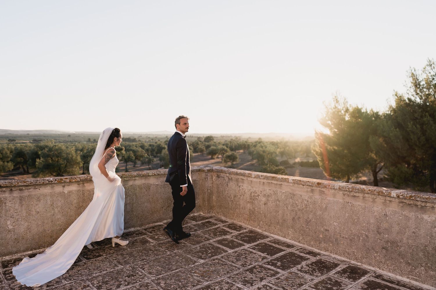 Wedding couple standing on stone terrace overlooking landscape at sunset.