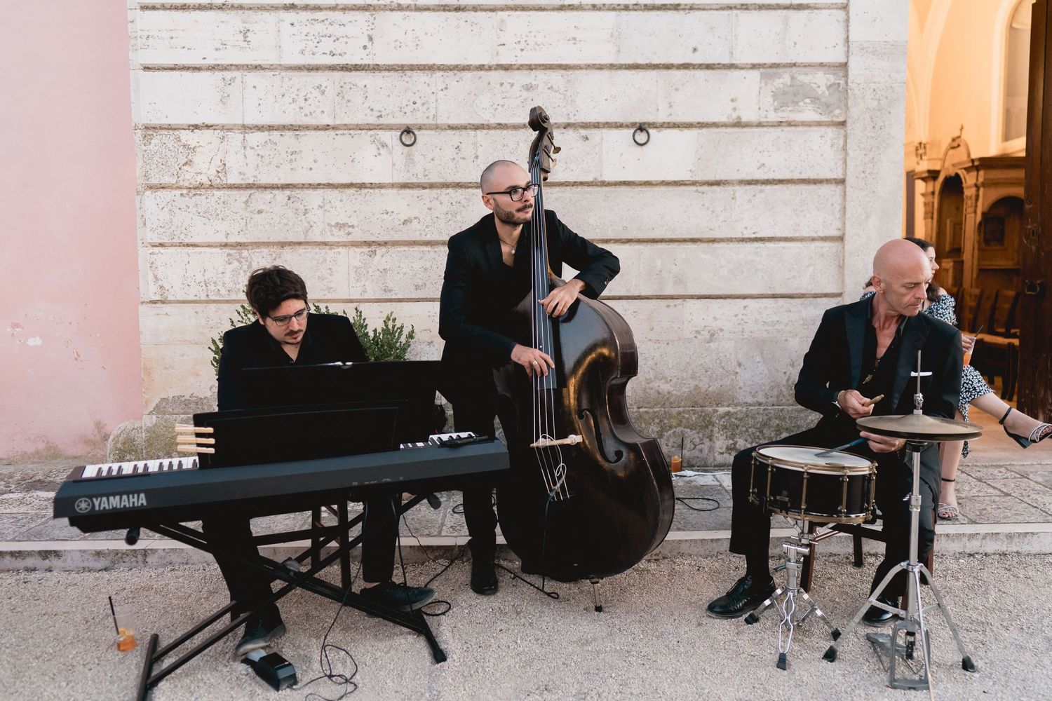 Three piece jazz band performs at outdoor wedding with keyboard, upright bass and drums against white wall.