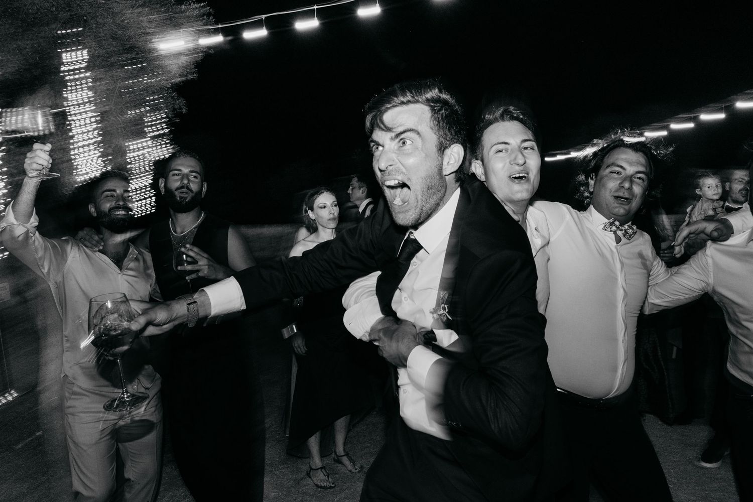 Black and white photo of a lively dance party with string lights overhead.