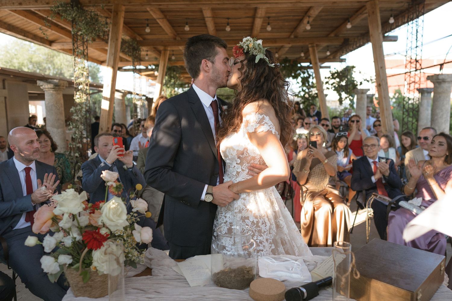 Celebration moment during an outdoor wedding ceremony under a wooden canopy with floral arrangements.