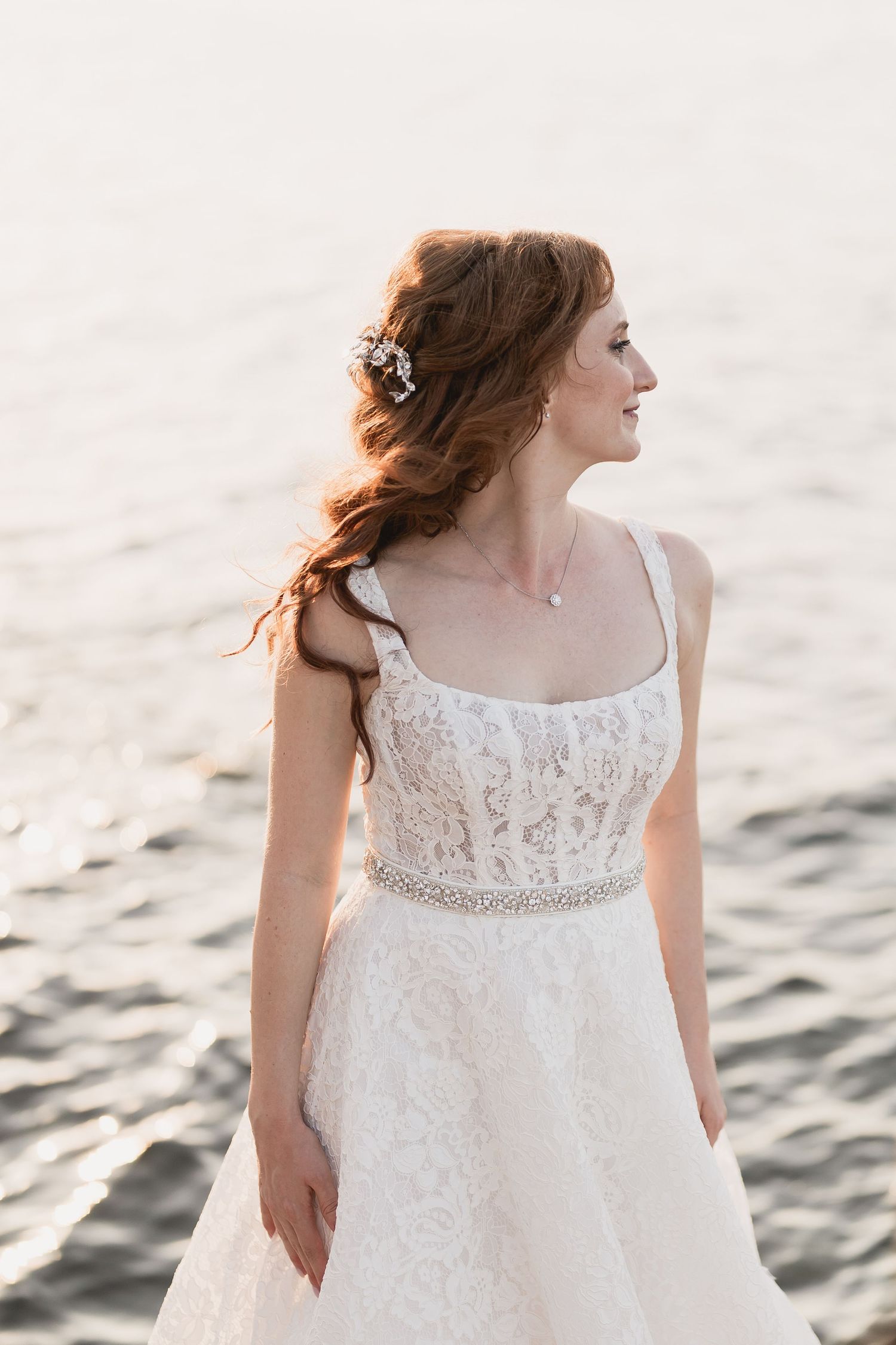 Elegant bride in lace wedding gown poses by the ocean at golden hour.