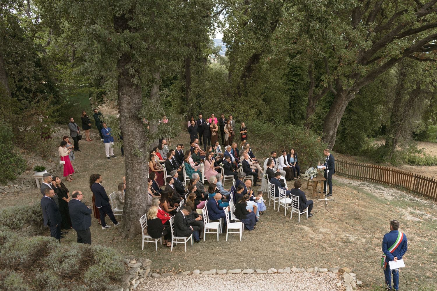 Wedding guests walk along a wooded path at an outdoor ceremony venue in a natural setting.