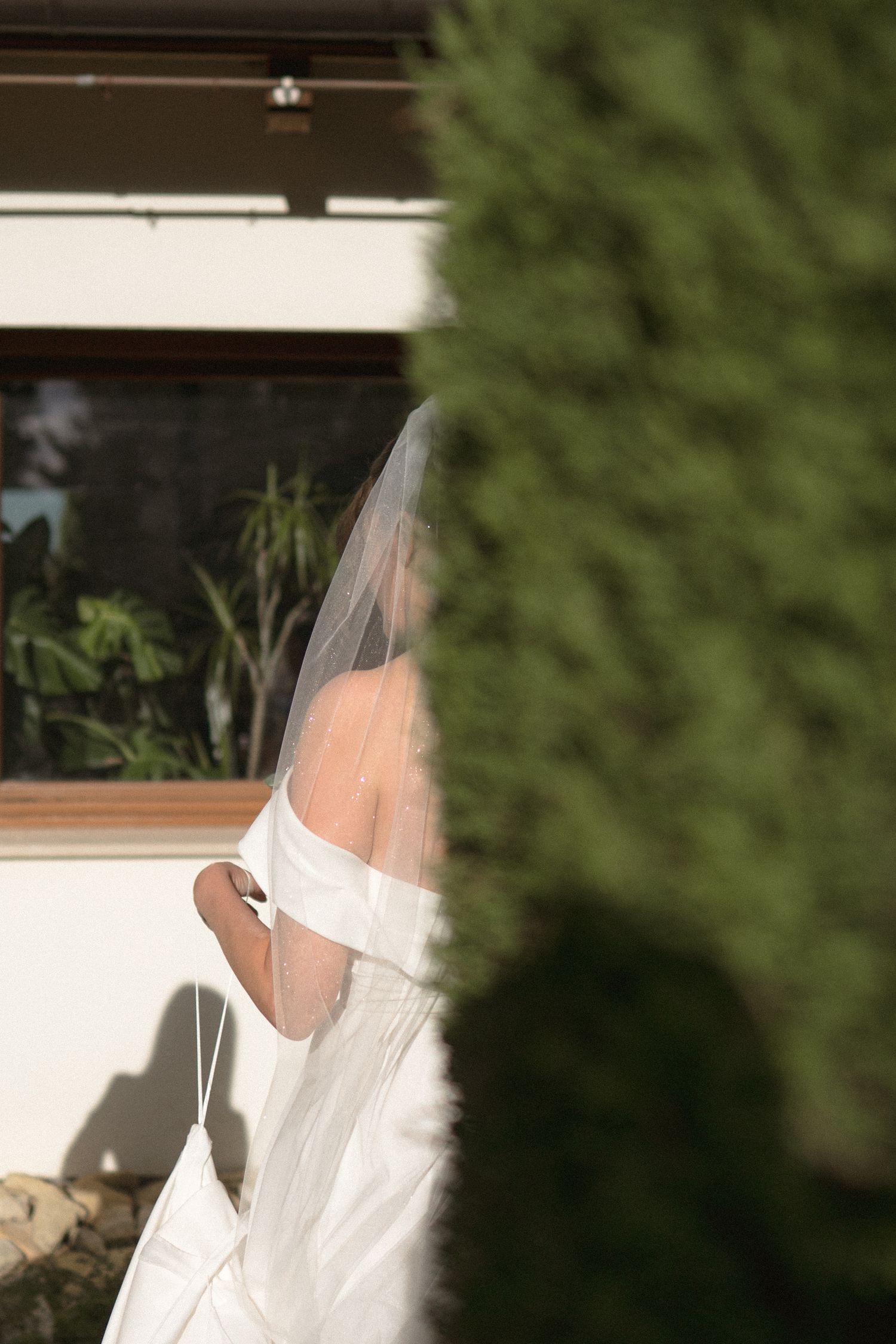 A bride in a white dress and veil stands in front of a window with natural light streaming in.
