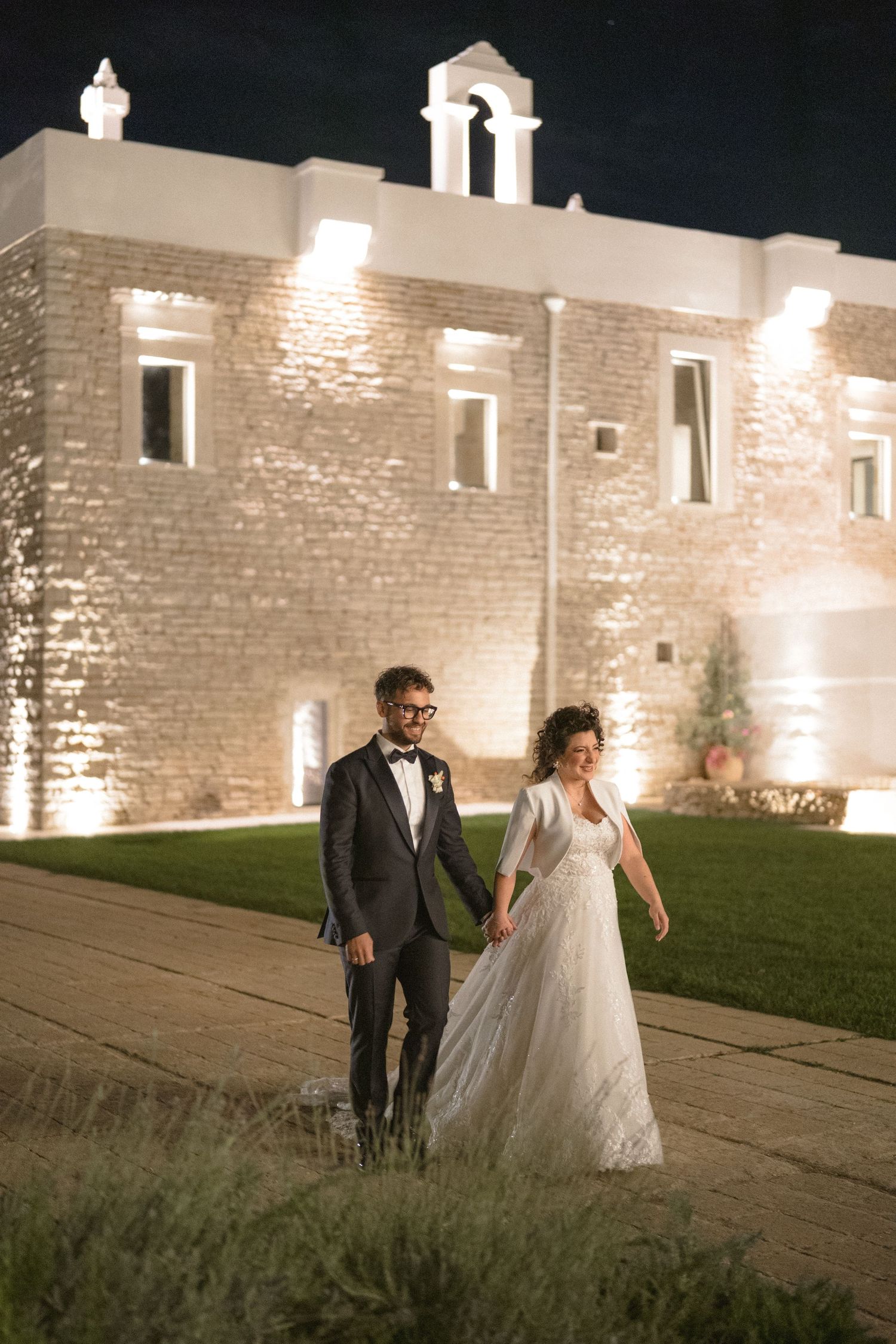 Newlywed couple holding hands outside an illuminated stone church building at night.