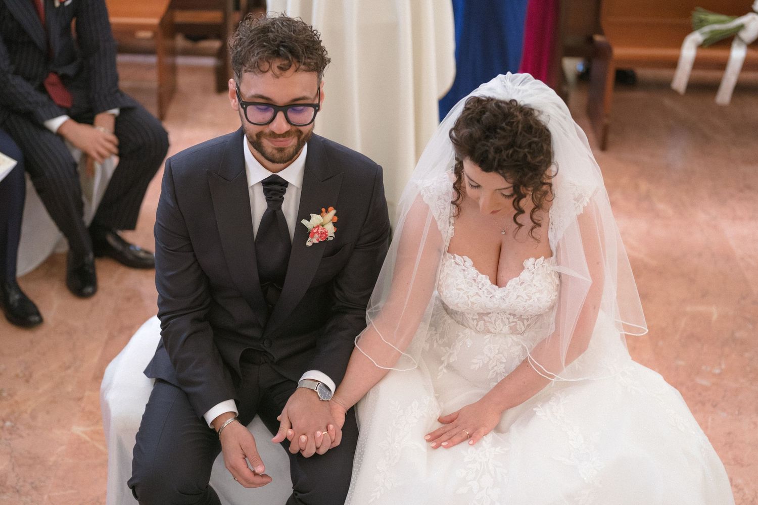 Bride in strapless lace gown and groom in navy suit hold hands during church wedding ceremony.