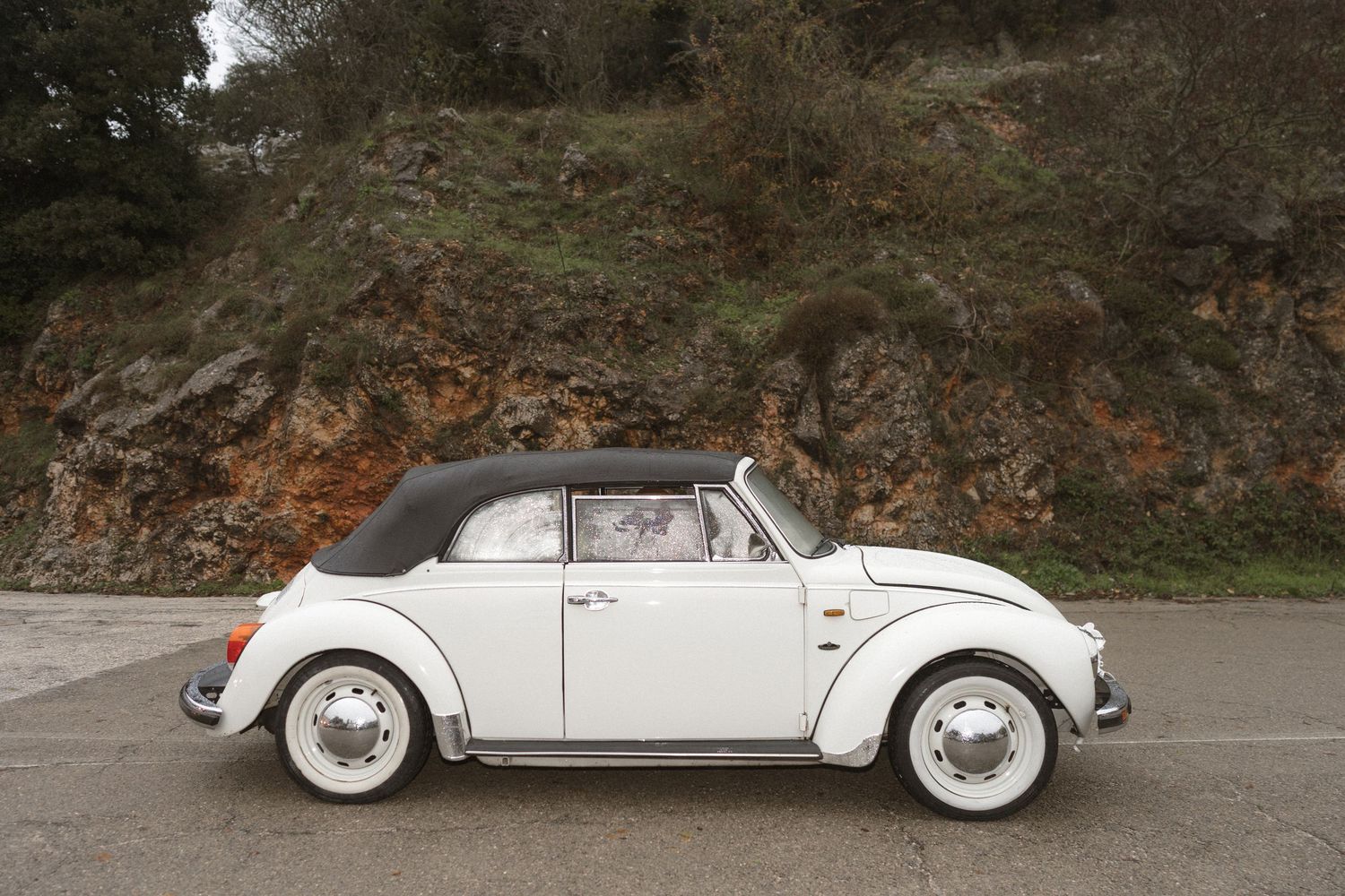 Classic white Volkswagen Beetle convertible parked on a road with rocky hillside background.