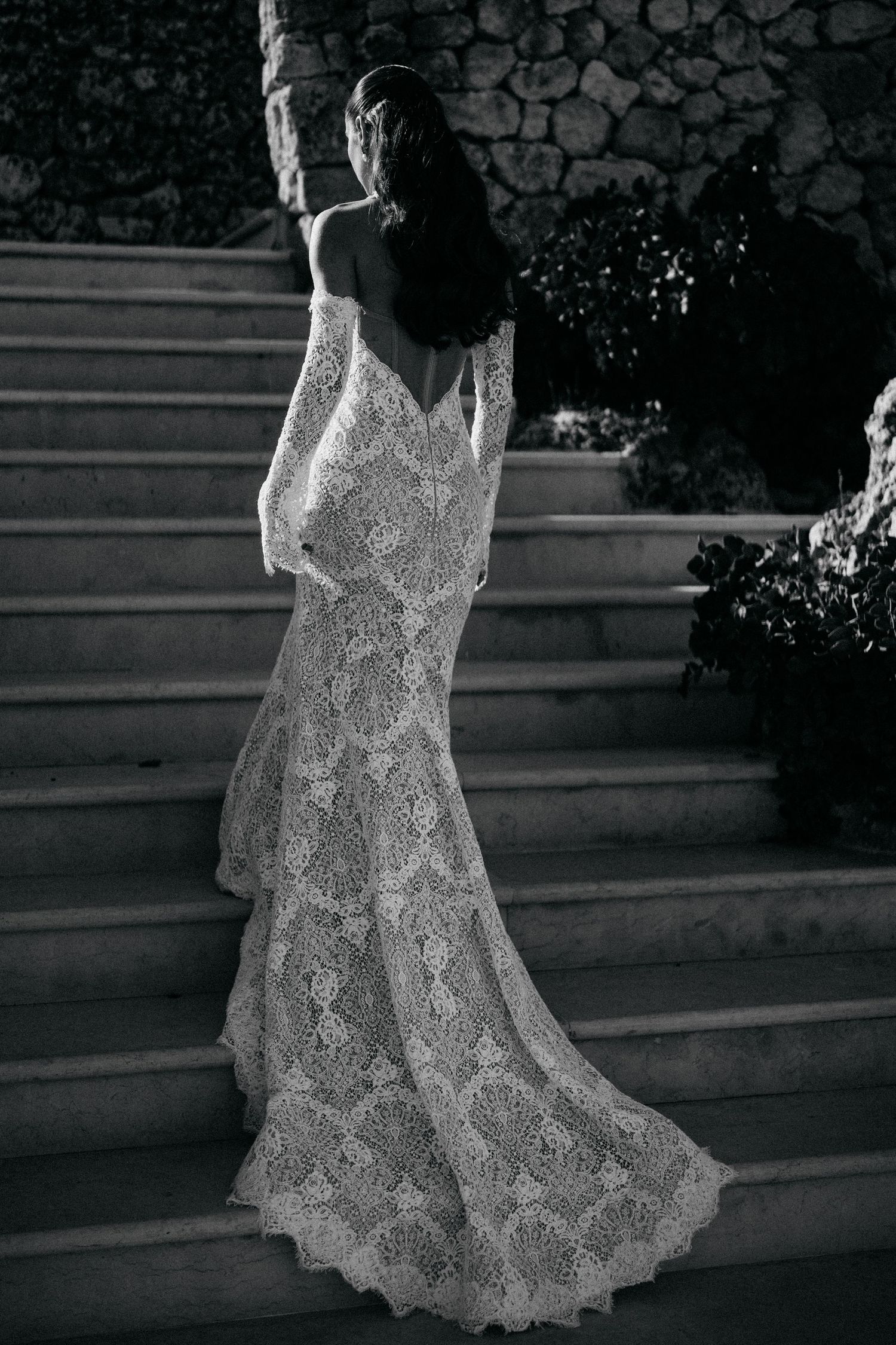 Black and white photo of a bride in a backless long-sleeve lace gown with a flowing train walking up stone steps.