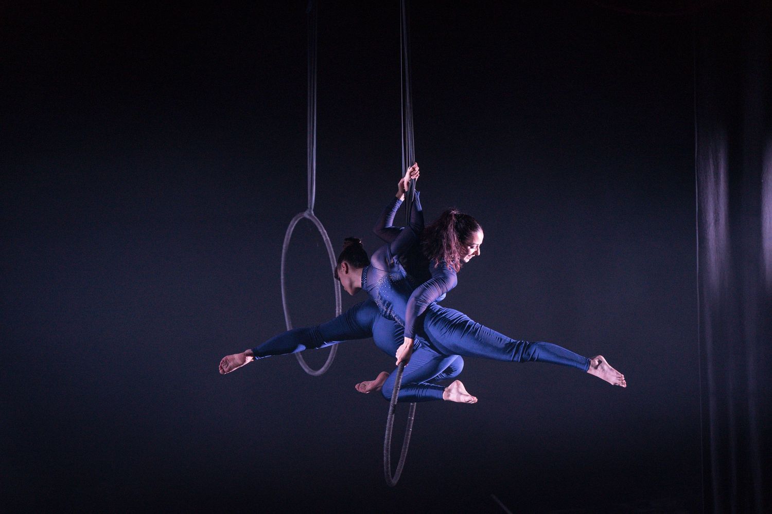 Aerial artist in blue costume performs dynamic pose on suspended hoop against dark background.