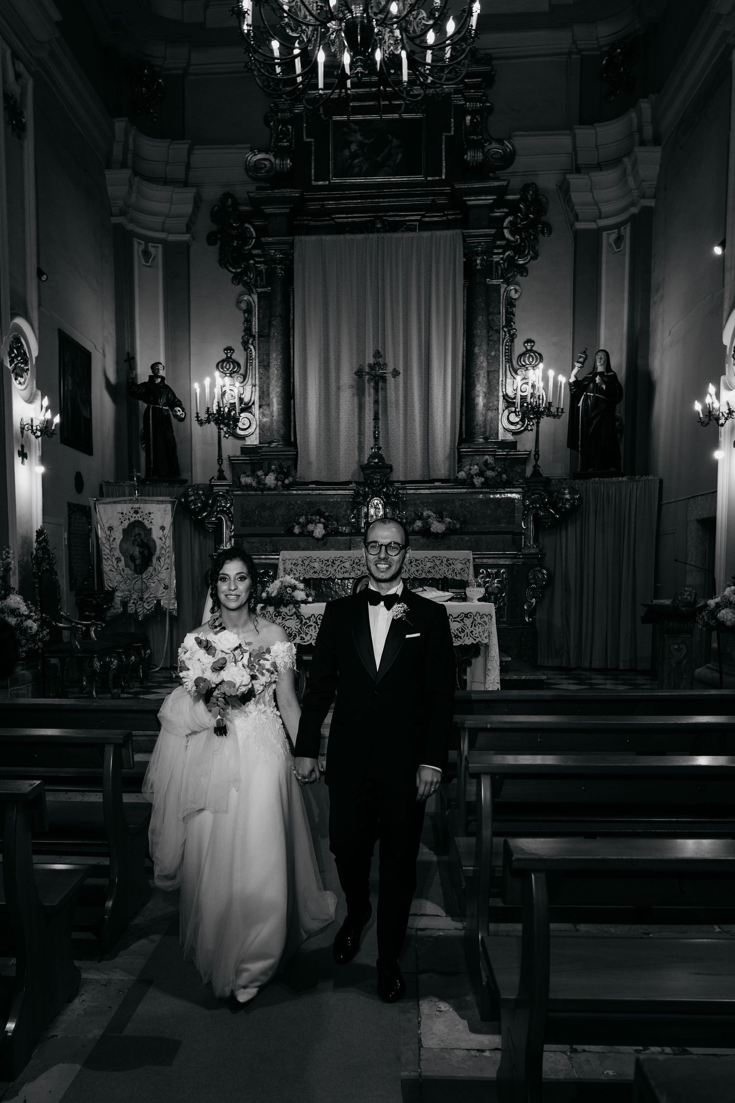 A black and white photo of a wedding ceremony in an ornate church with chandeliers and decorative altar.