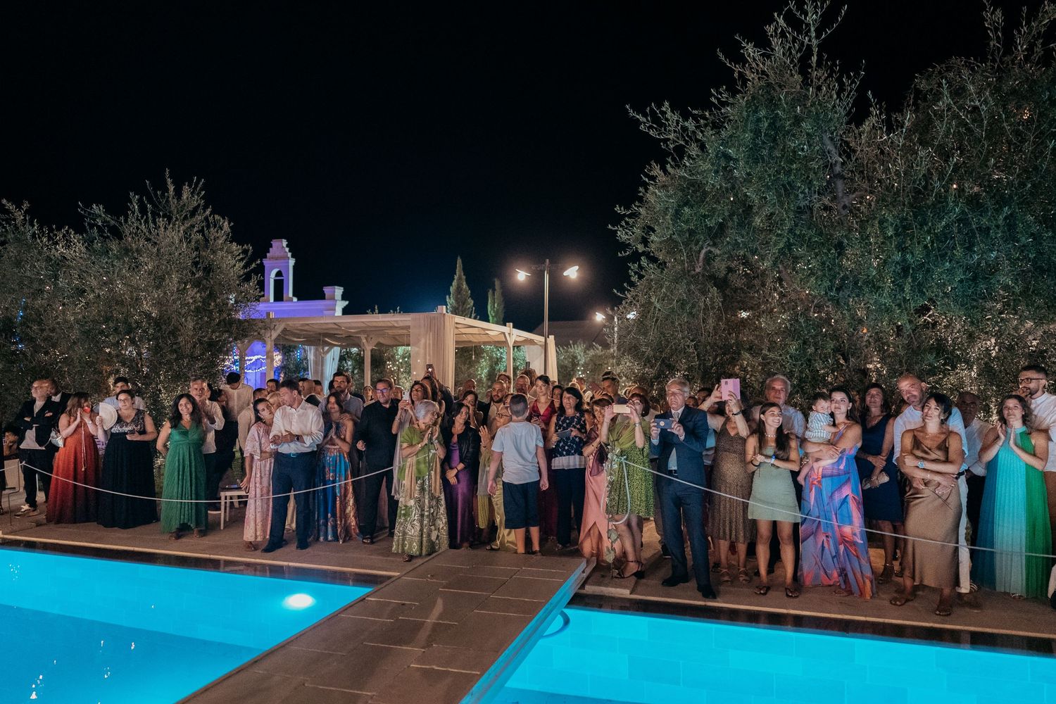 Wedding guests gather around illuminated pool during evening outdoor reception celebration.