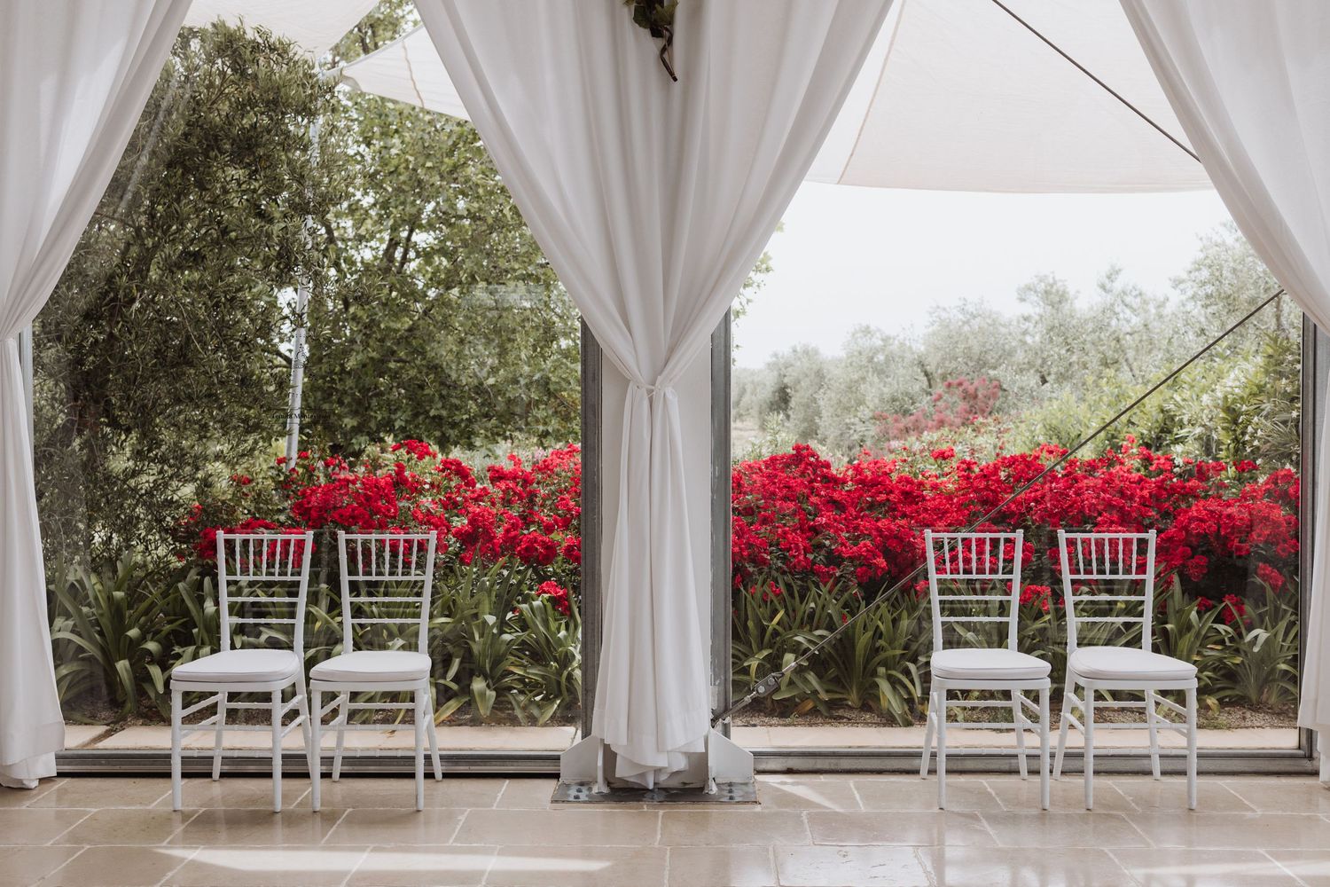 White chairs set up under flowing white drapes with vibrant red bougainvillea in the background.