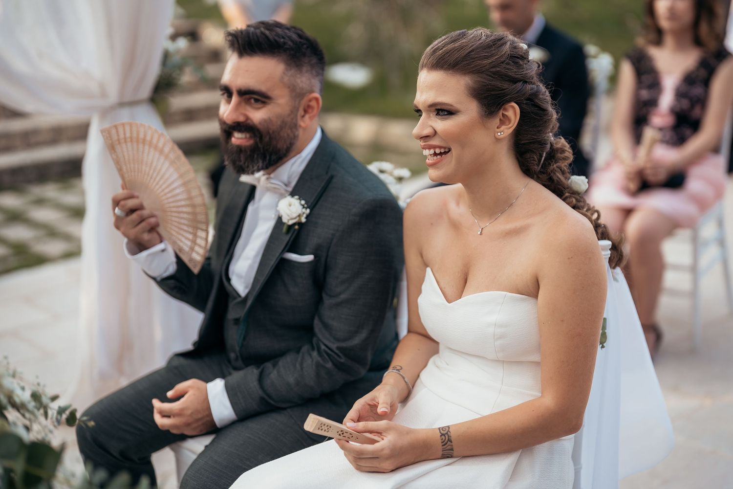 A couple sits together during their outdoor wedding ceremony sharing a light moment.
