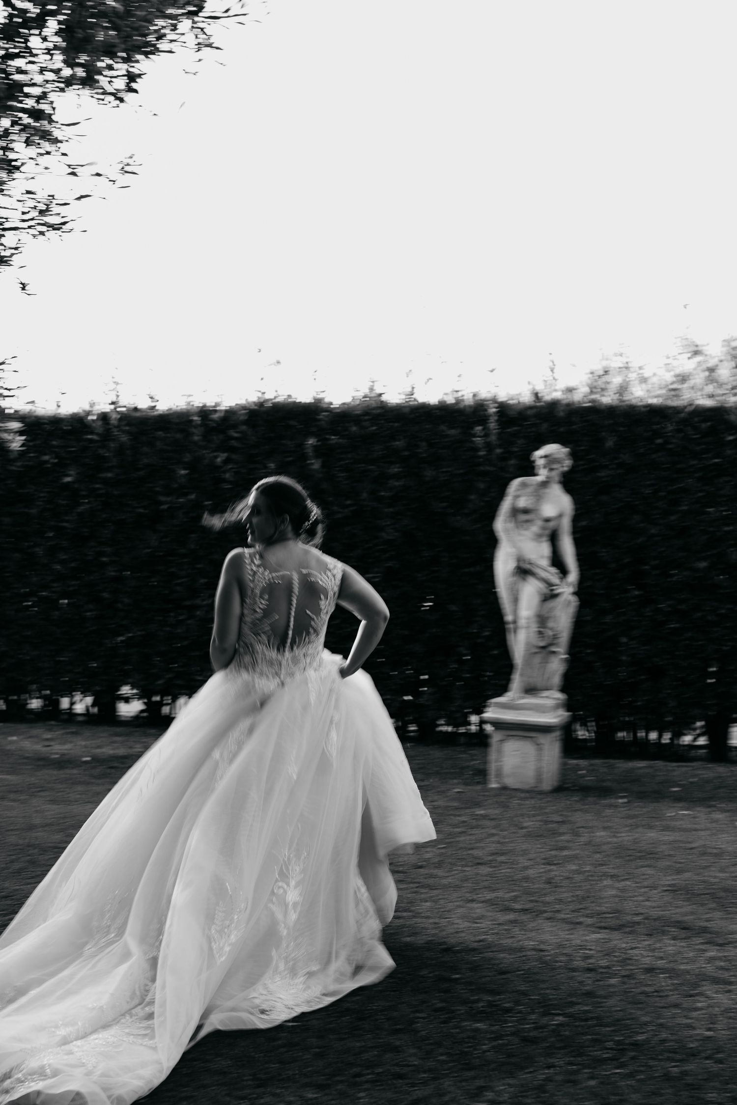A dramatic black and white photo of a bride in a ballgown looking at a classical statue in a formal garden.