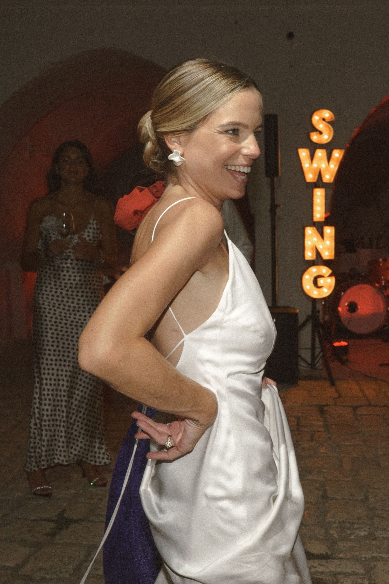 Woman in white dress dancing joyfully at reception with illuminated 'SWING' sign in background.