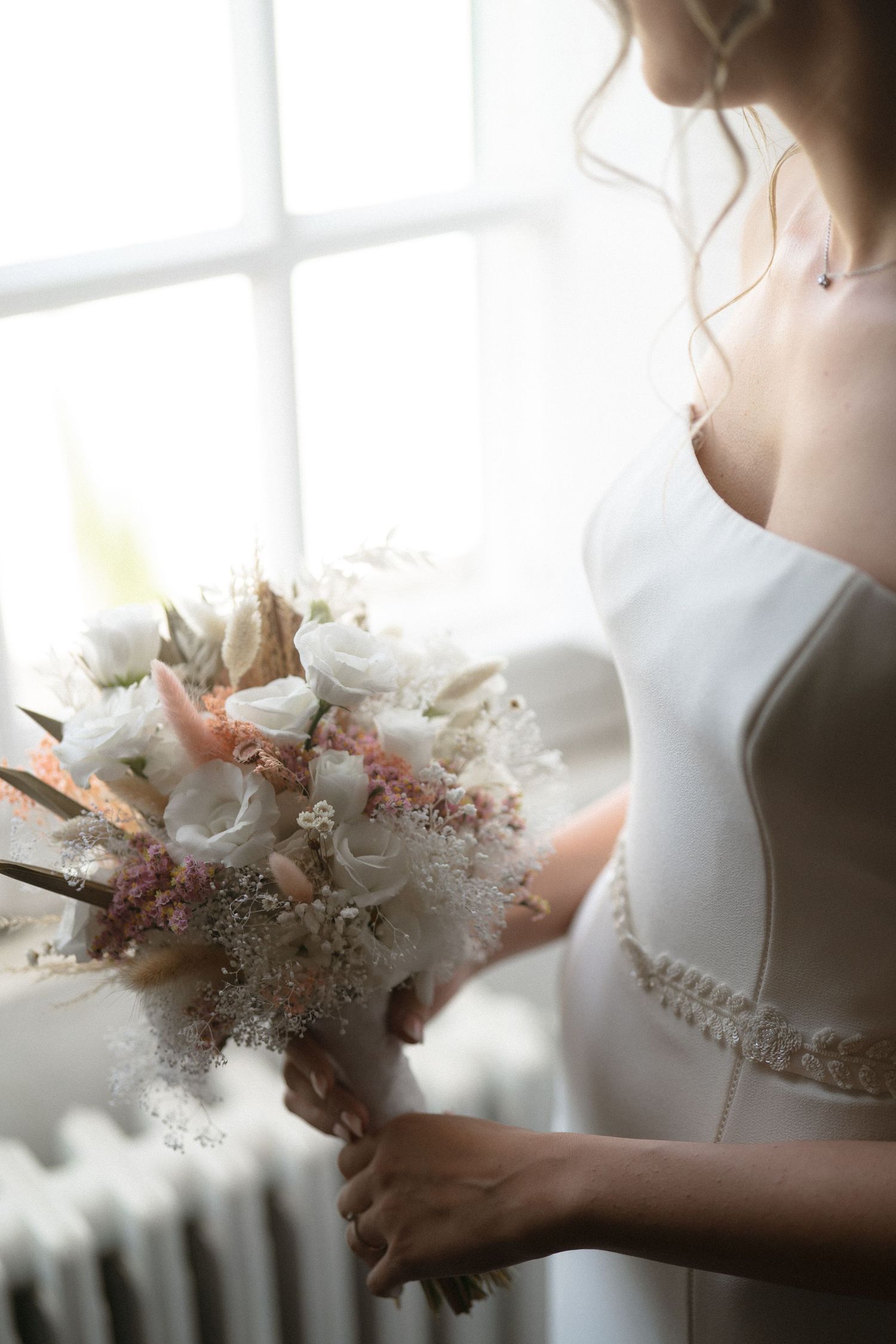 Delicate white and pink bridal bouquet held near sunlit window.