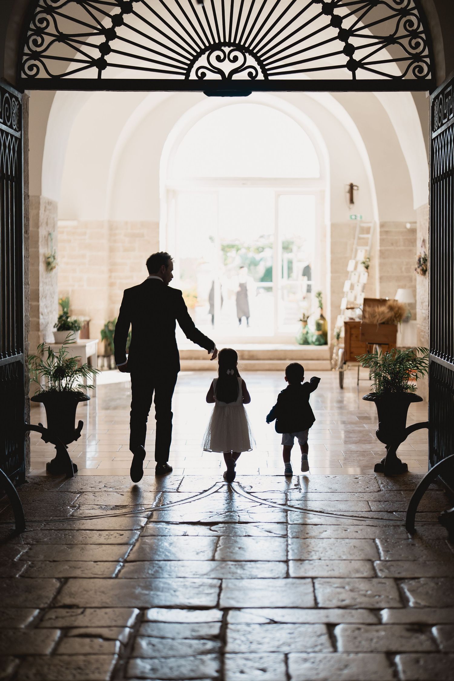 Silhouettes of figures walking through an elegant arched doorway with ornate iron details and stone flooring.
