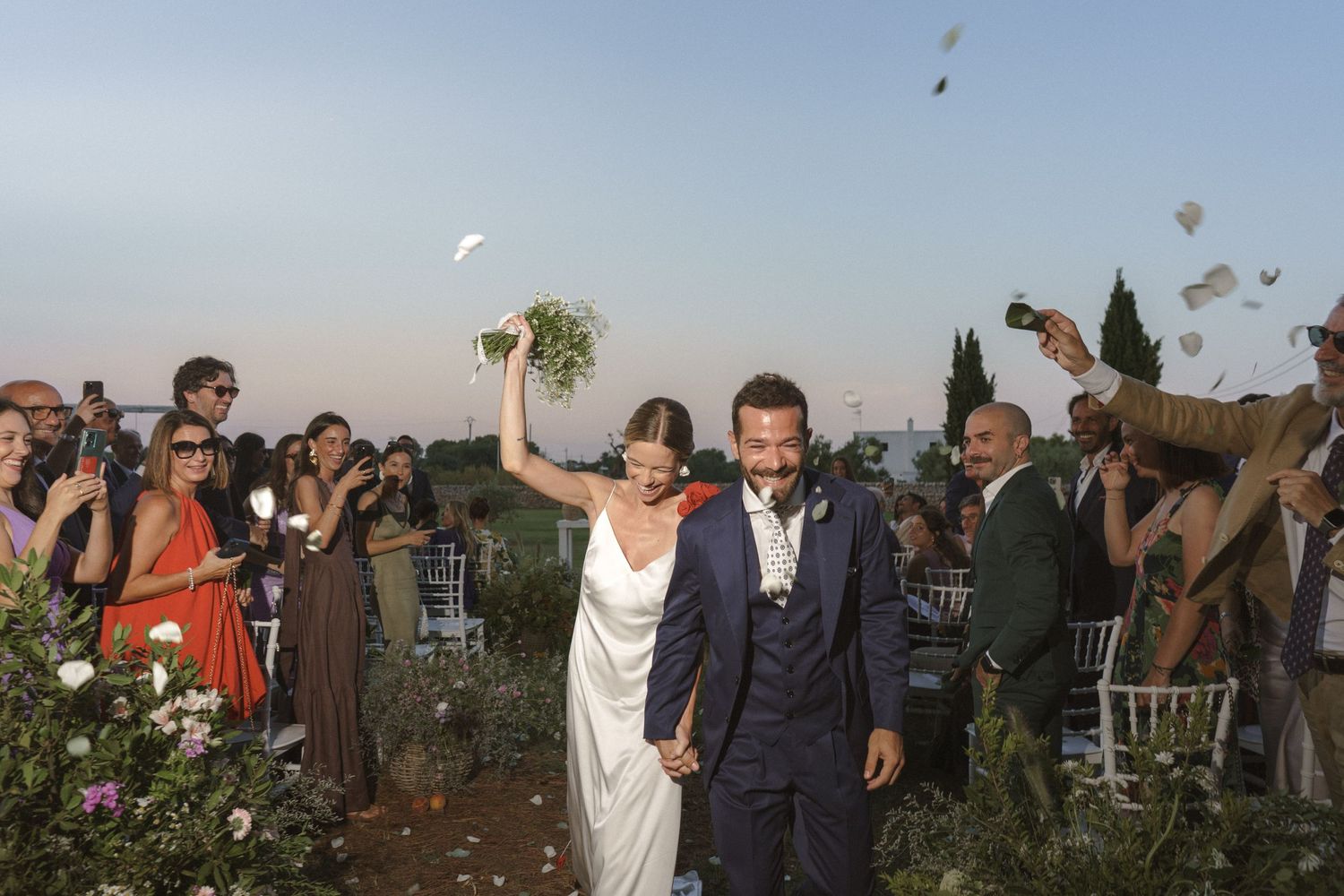 Wedding guests throwing confetti as newlyweds walk down aisle during outdoor celebration.