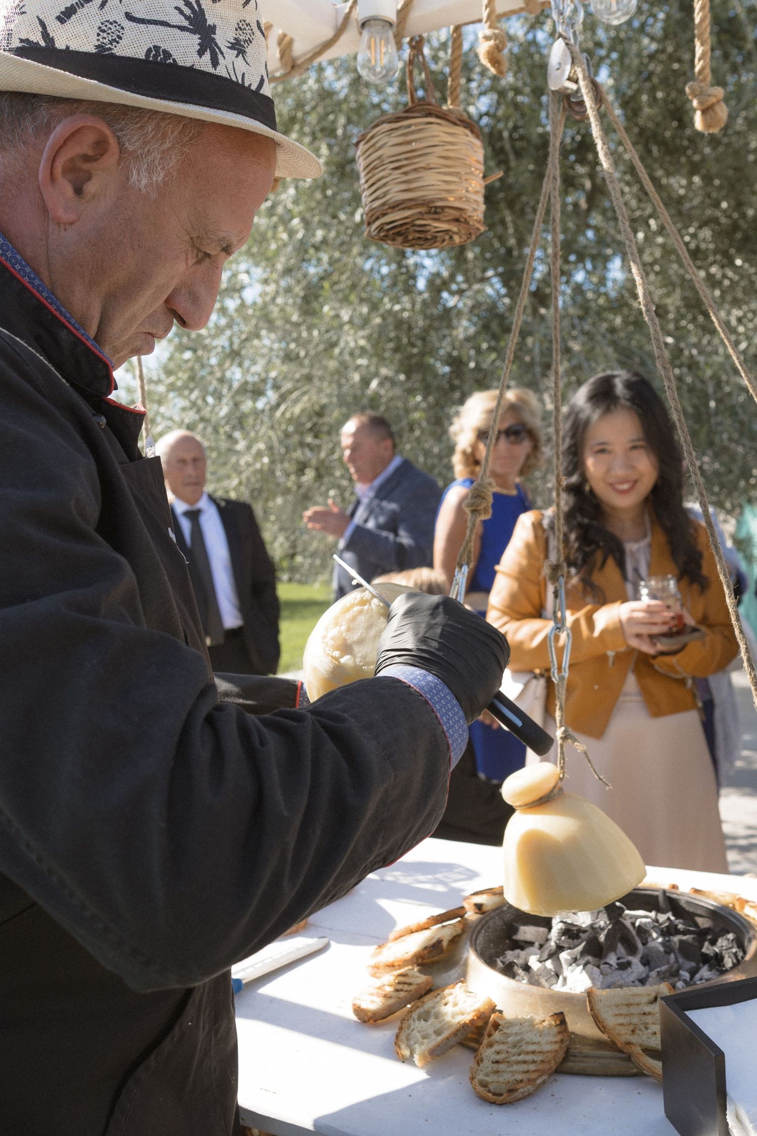 Man in hat carves fresh cheese at outdoor wedding reception as guests look on.