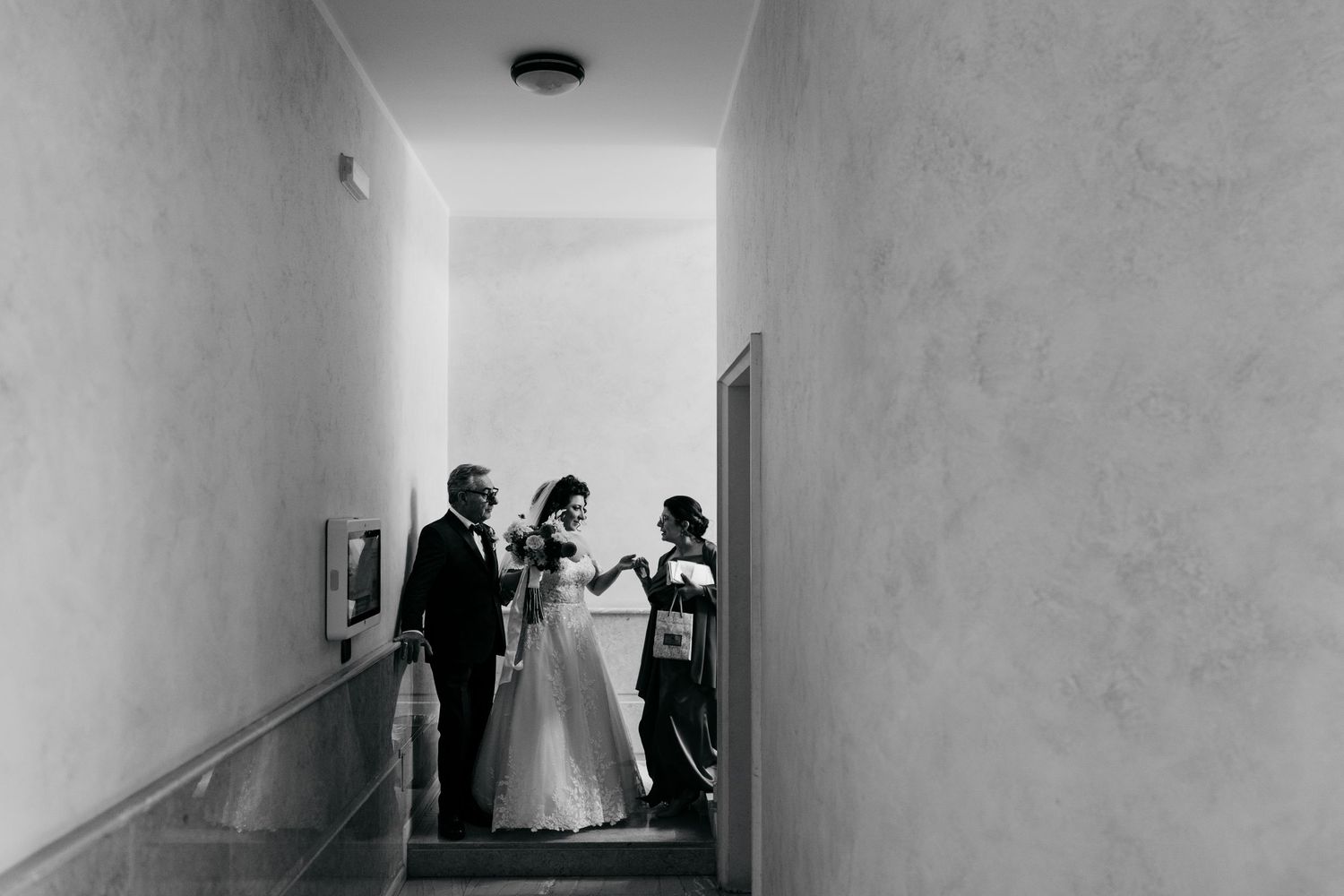 Black and white wedding photo of bride in gown with groom and guest in a hallway.