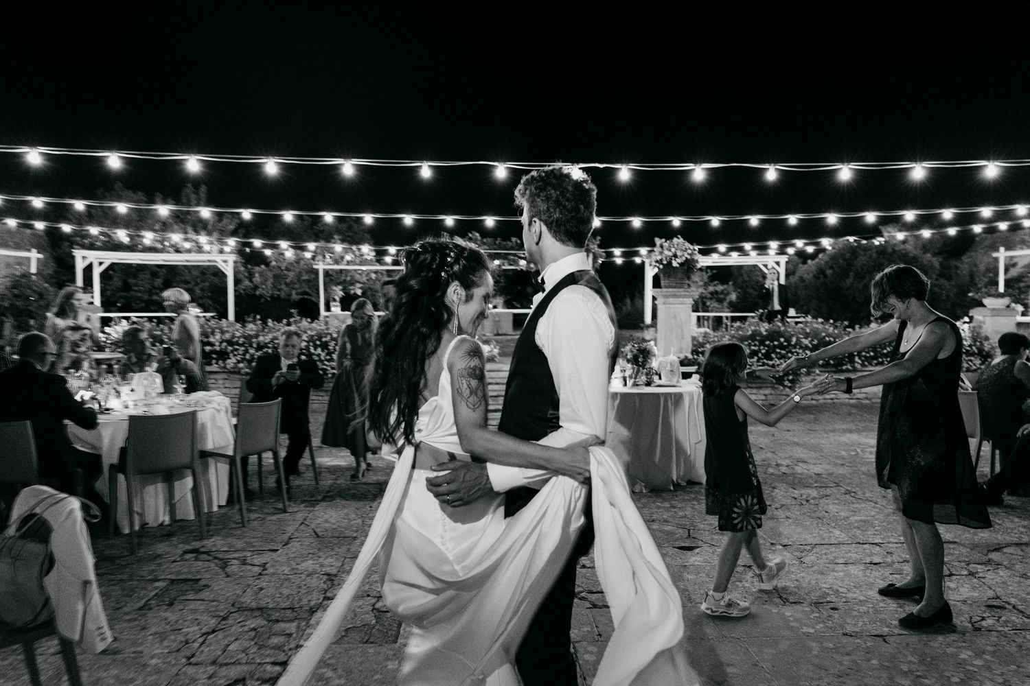 A black and white photo of a wedding reception under string lights with guests dancing at an outdoor venue.