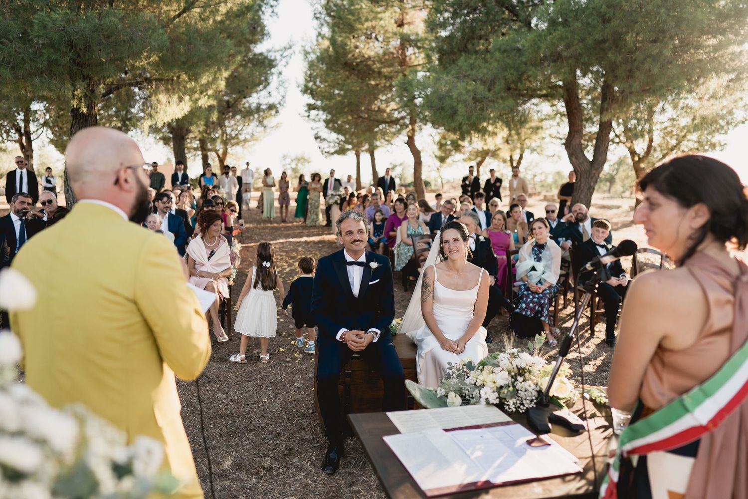 An outdoor wedding ceremony takes place in a pine forest with guests gathered to witness the occasion.