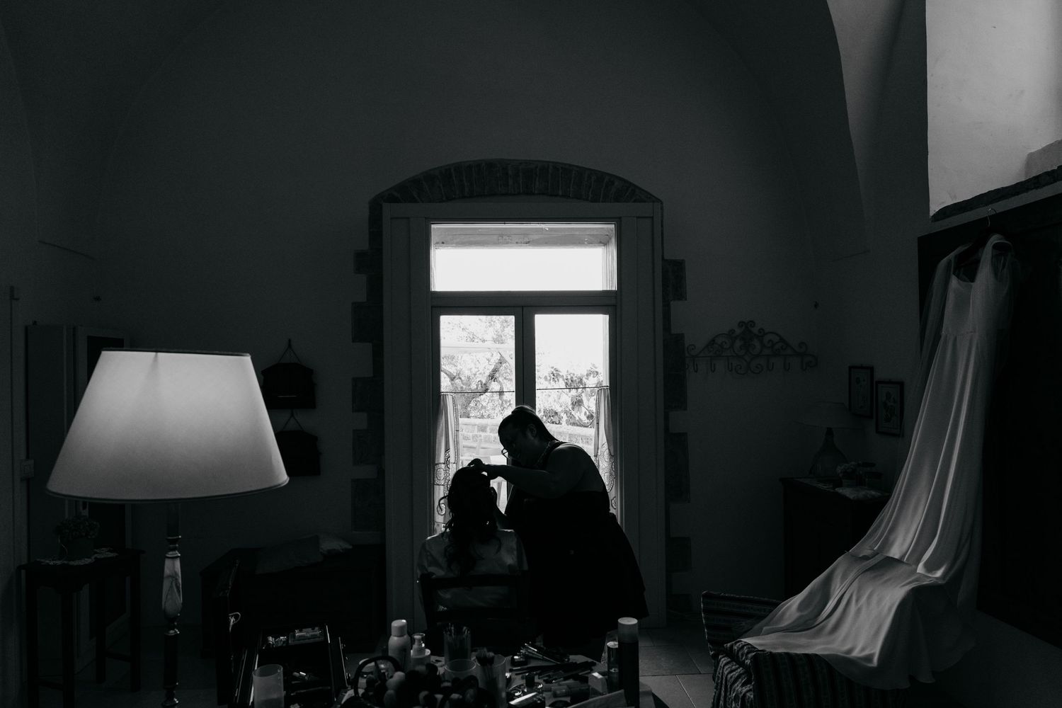 Dark silhouettes against a bright window in a dimly lit room with a table lamp creating moody ambiance.