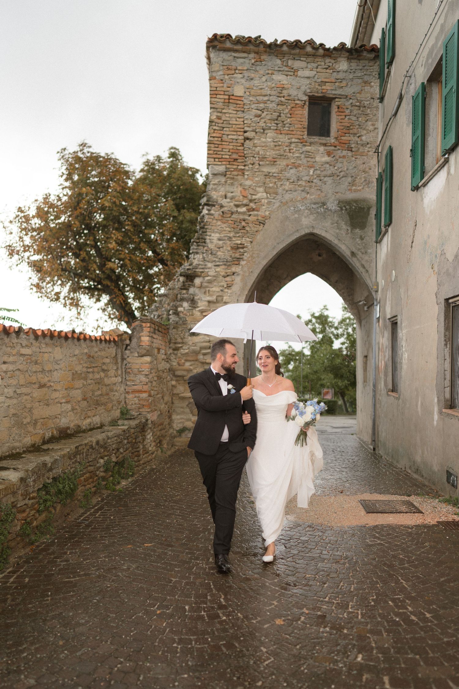 Couple walks along a cobblestone path beside historic stone buildings holding a white umbrella.