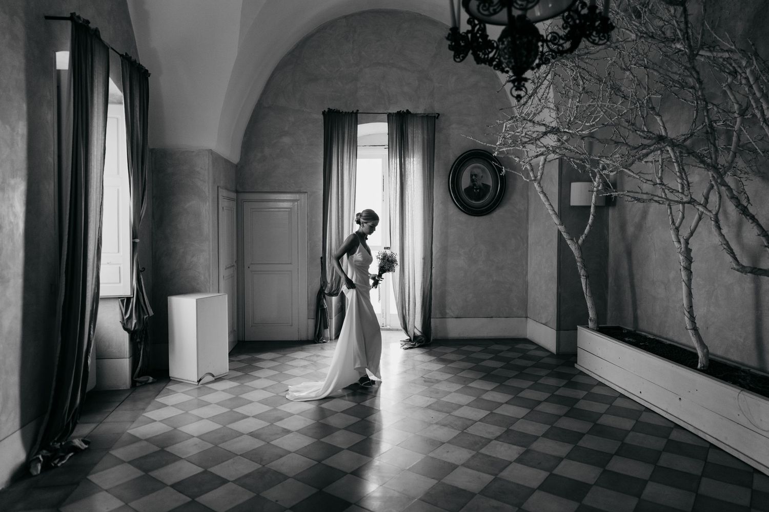 A figure stands in dramatic light streaming through windows in an elegant hallway with checkered floors and vaulted ceilings.