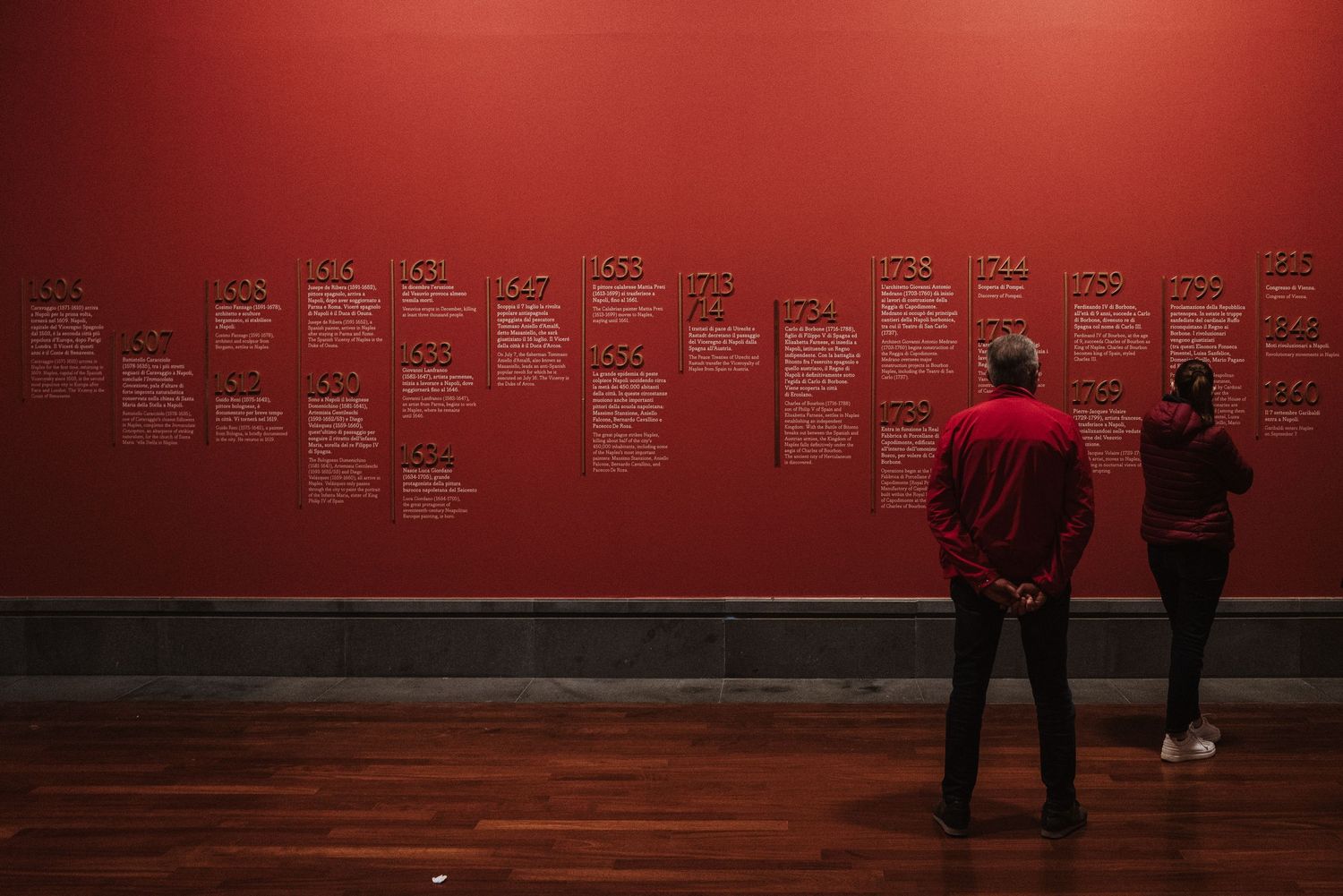 Two silhouettes observe red wall displays in a dimly lit museum or gallery space.
