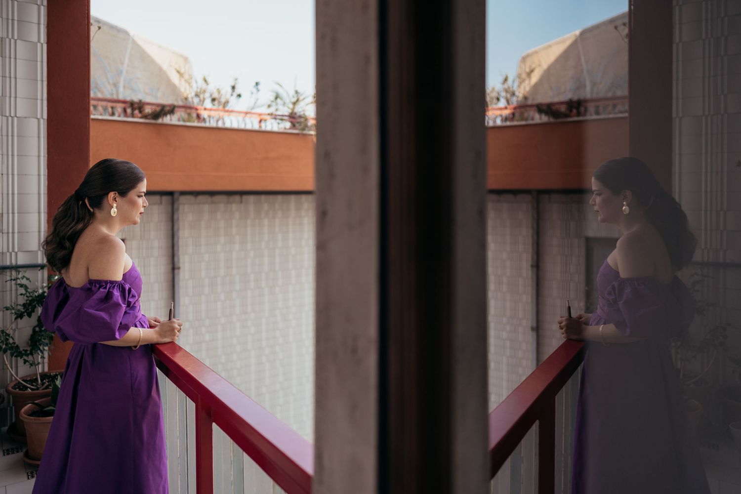 A person in a purple dress stands on a balcony with their reflection visible in the window.