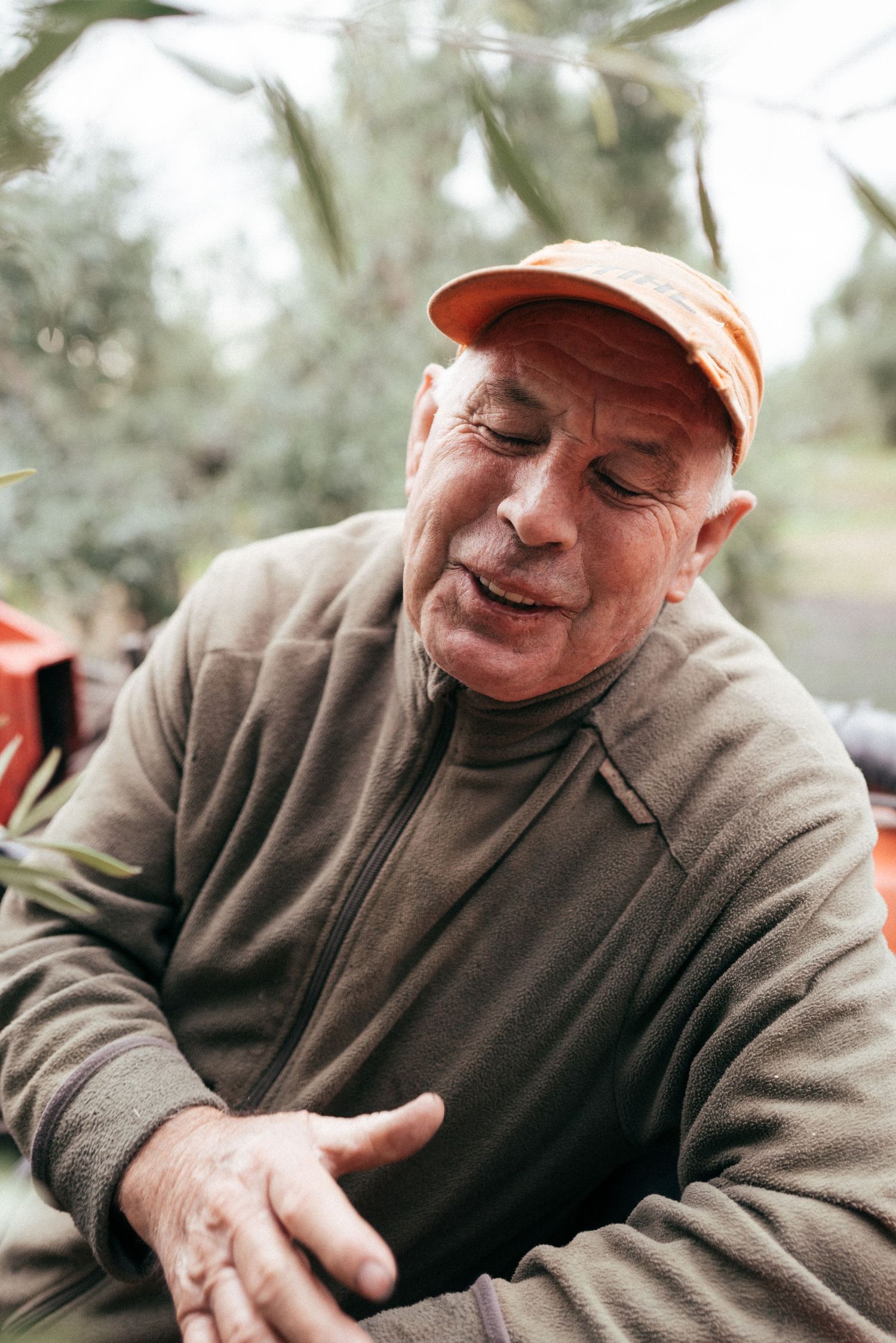 Someone in an orange cap and brown sweater sits outdoors with a relaxed expression on their face.