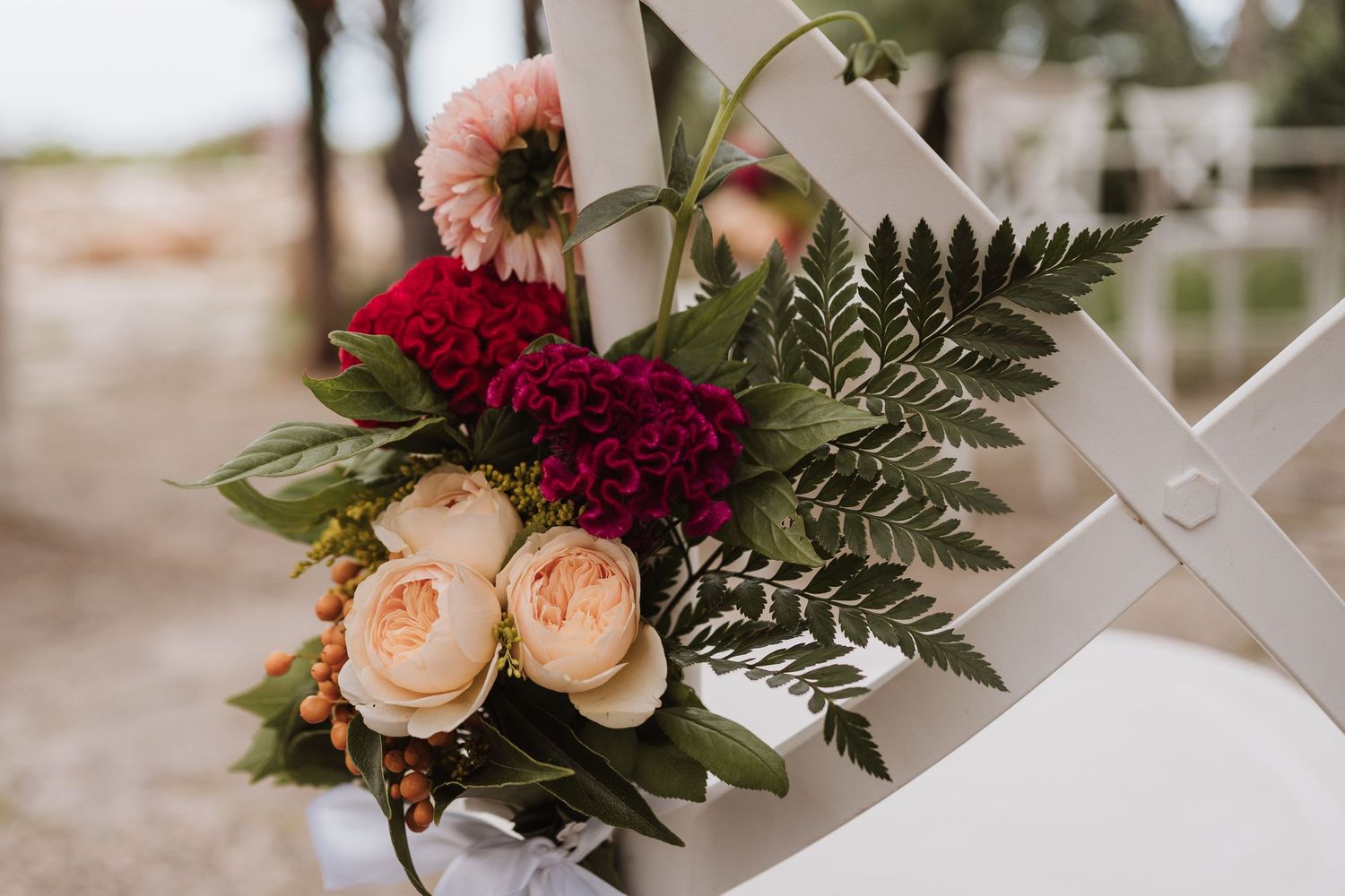 Wedding floral arrangement with pink roses, red flowers, and ferns decorating a white chair.