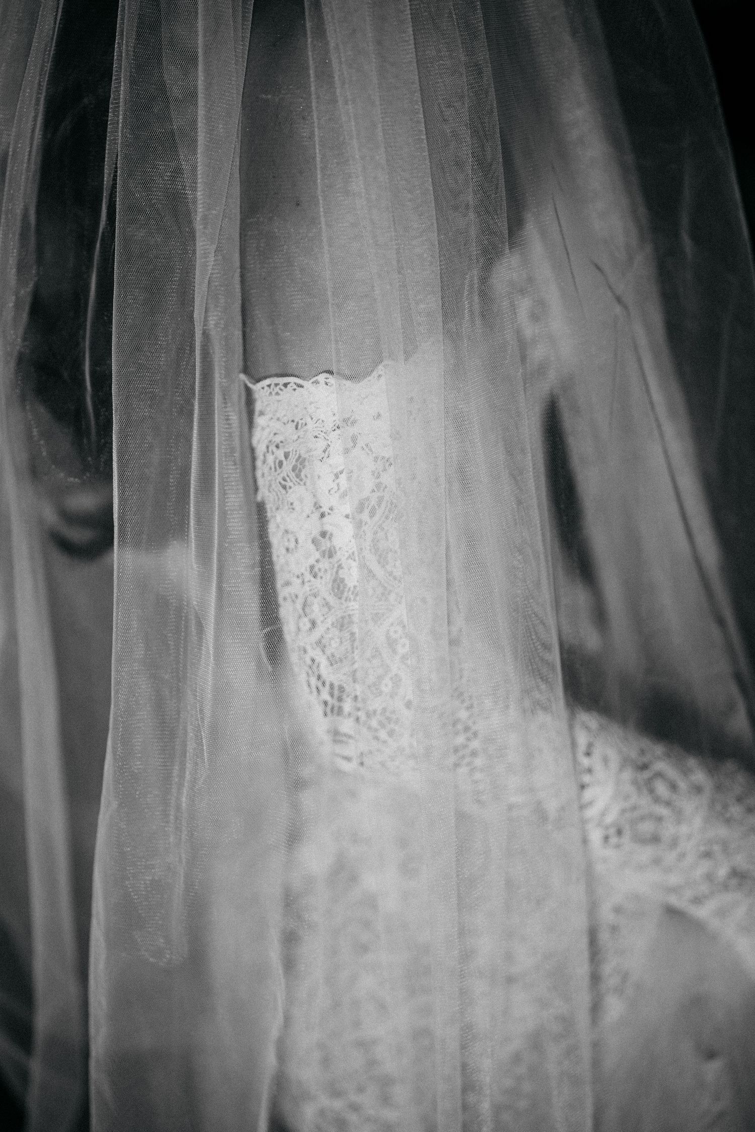 Black and white close-up of a bride in a lace wedding dress seen through a soft, sheer tulle veil.