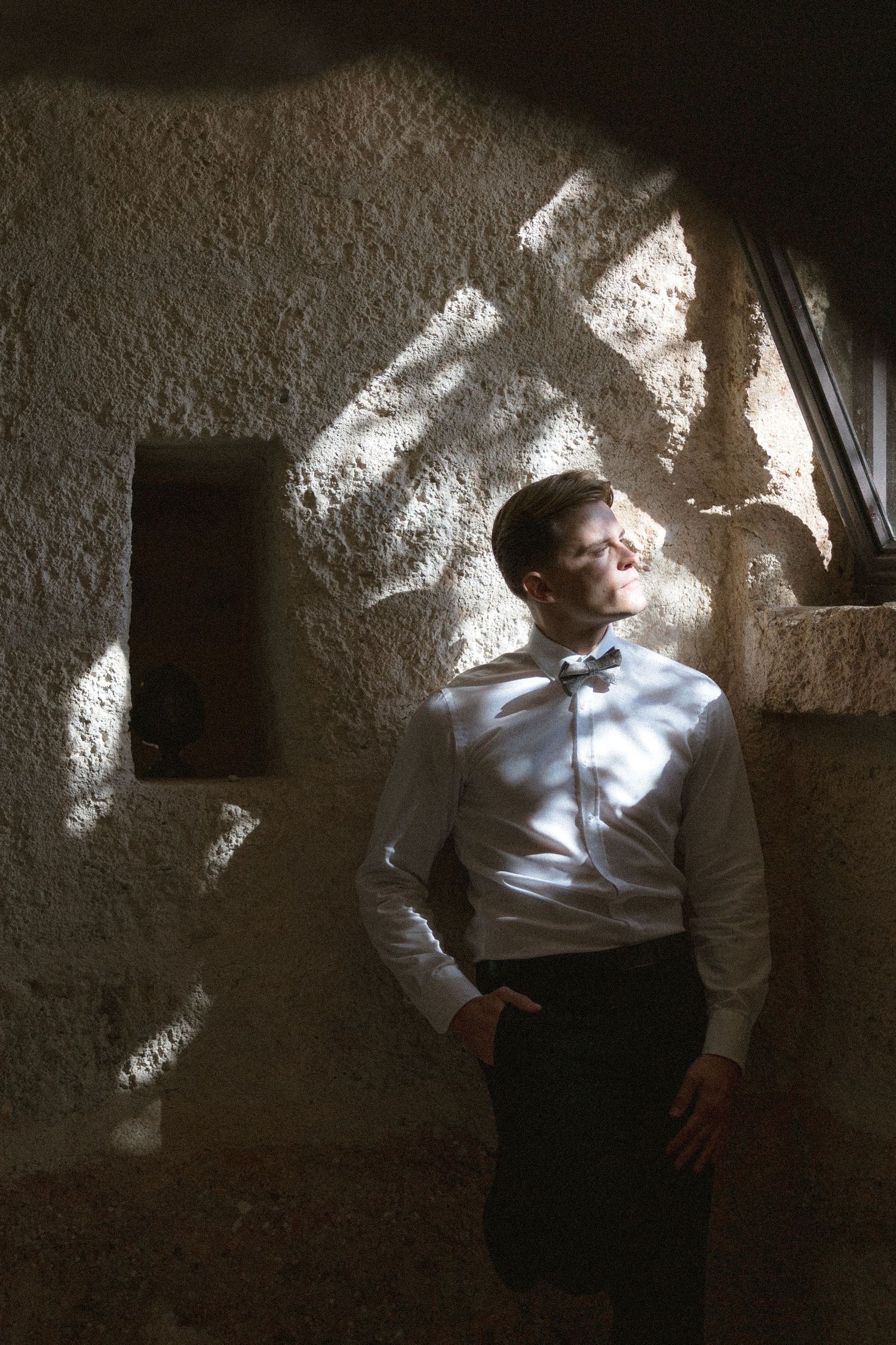 Groom in a white shirt and bow tie leans against a rustic stone wall, lit by dappled sunlight and shadows.