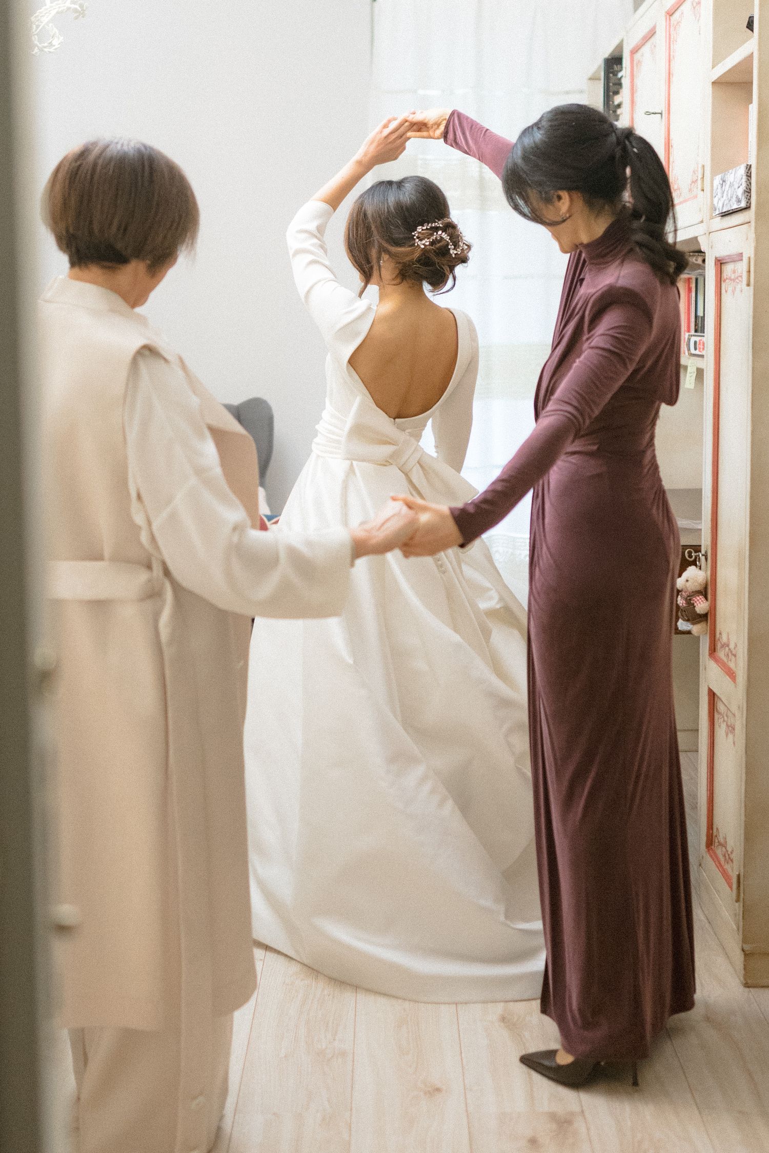 Bride in elegant backless white gown being assisted by two women while getting ready for wedding.