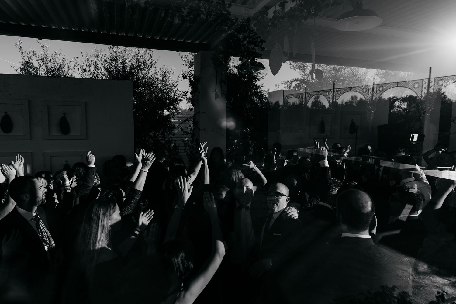 Black and white photo of wedding guests dancing with hands raised at an energetic outdoor reception.
