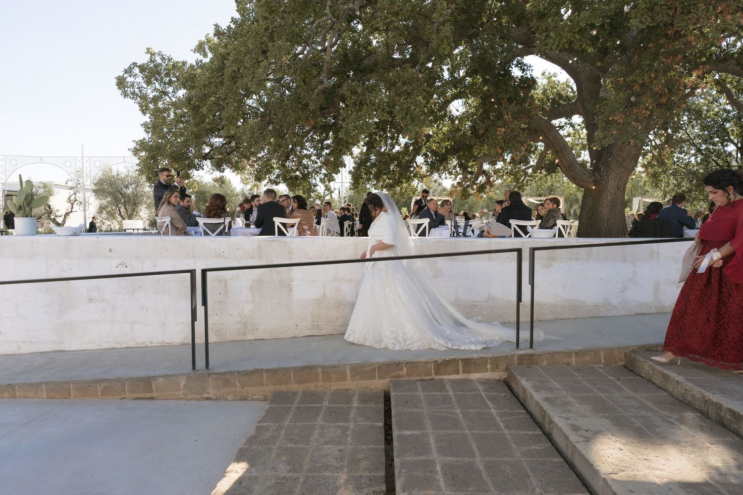Bride in flowing white gown walking along a terrace at an outdoor wedding ceremony.