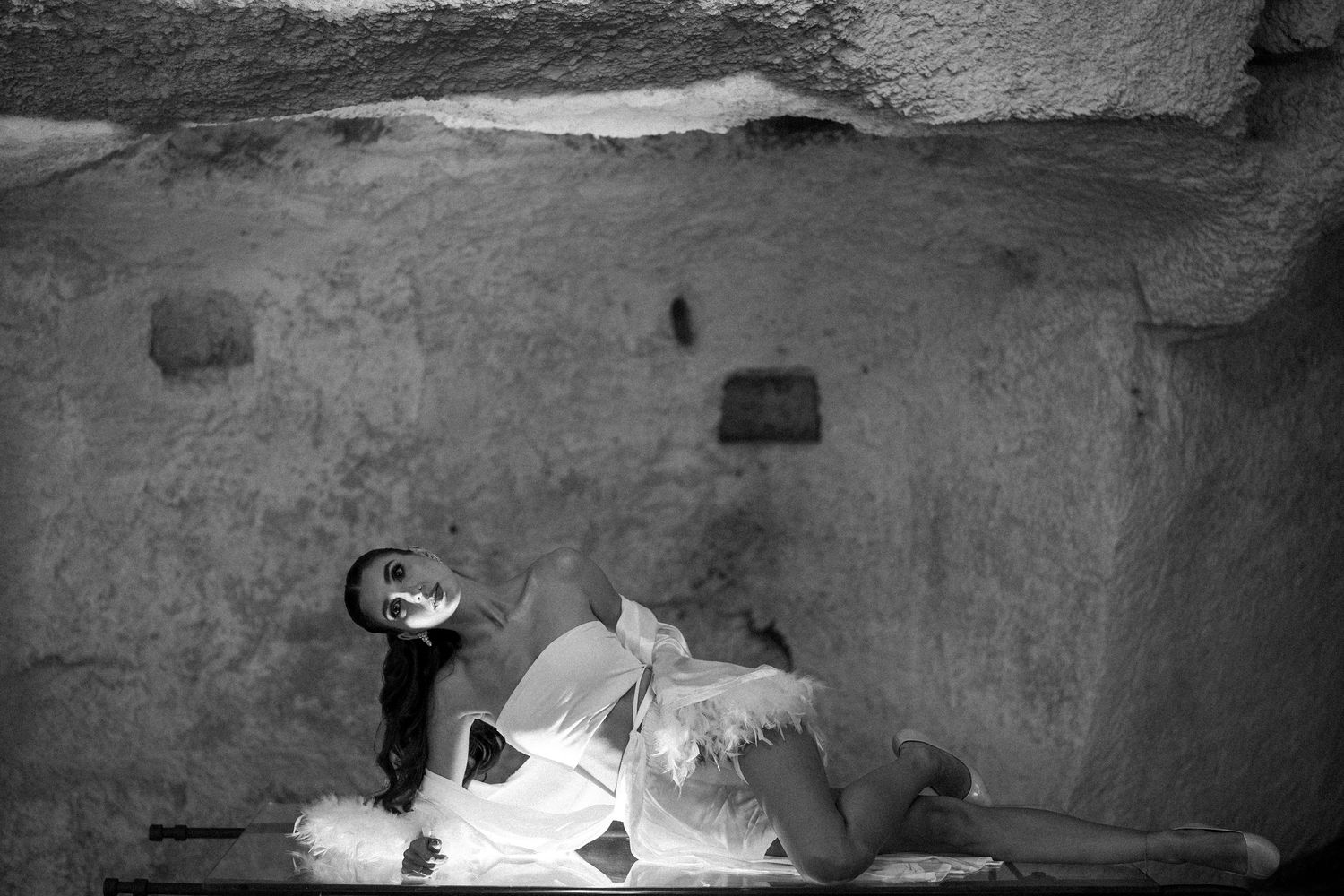 Black and white photo of a woman in a flowing white dress reclining on the floor inside a rough-hewn stone cave room.