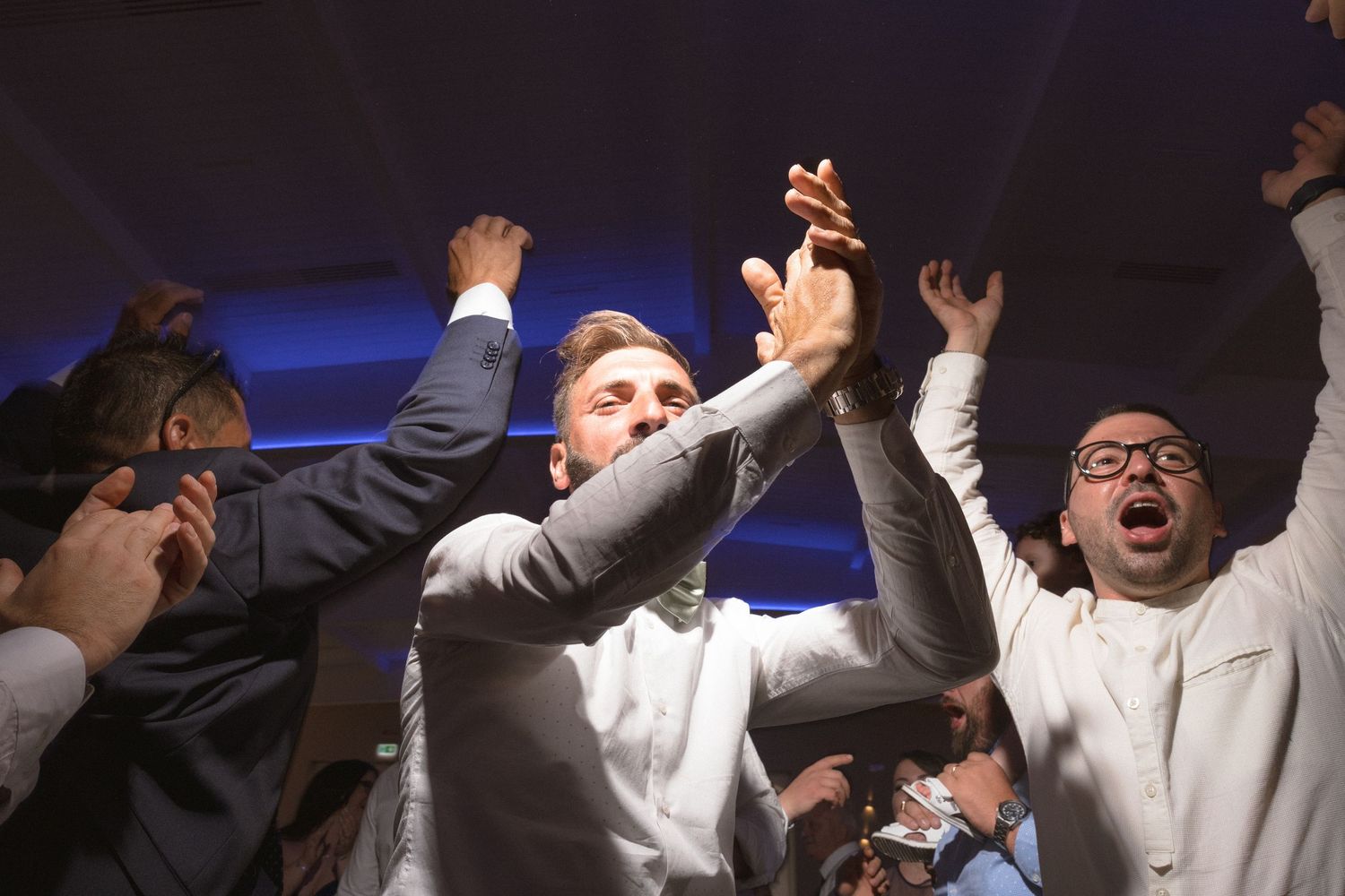 Wedding guests celebrate with raised arms and excited expressions on the dance floor.