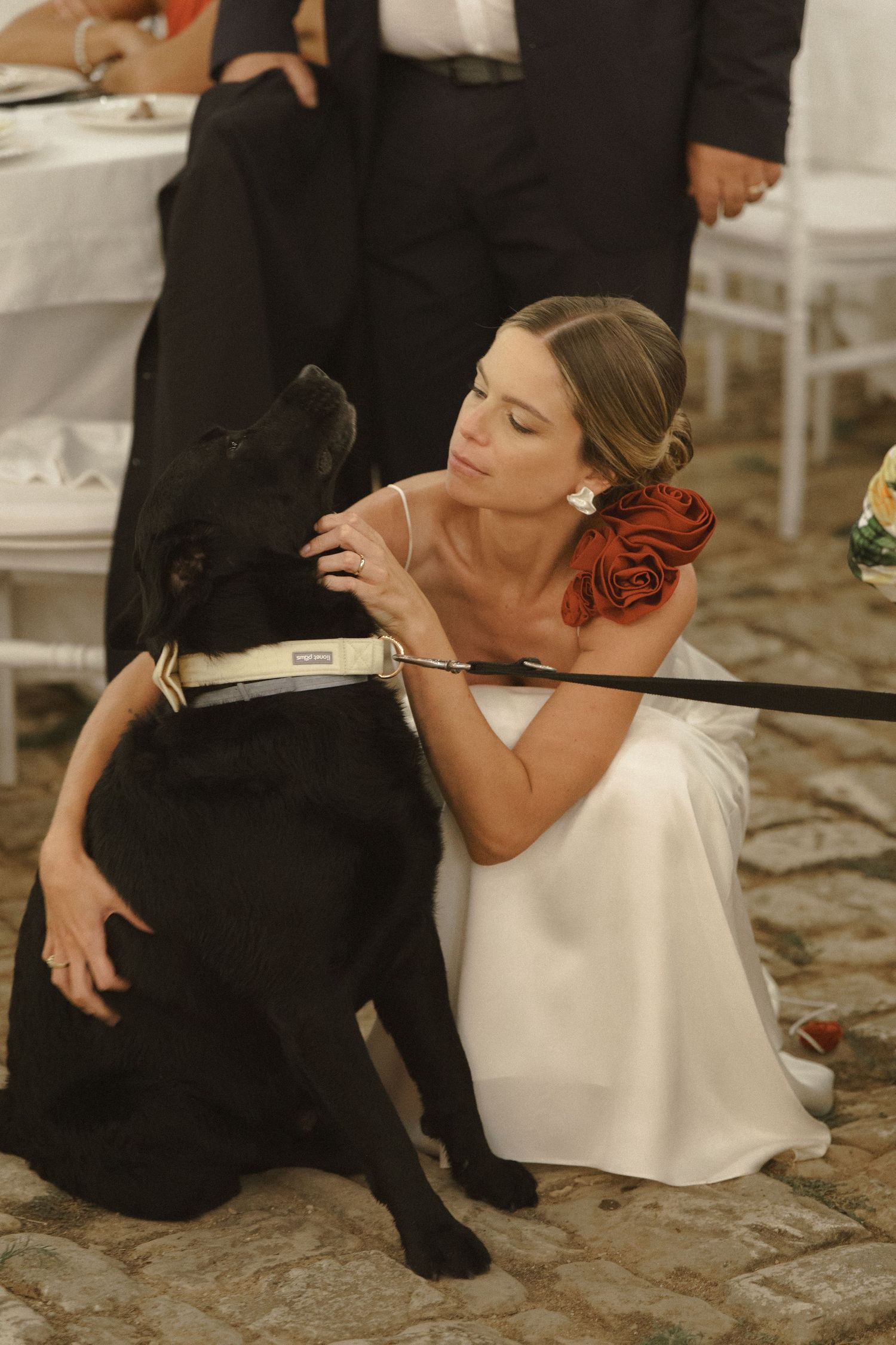 A person in a white wedding dress kneels down to pet a large black dog at an indoor celebration.