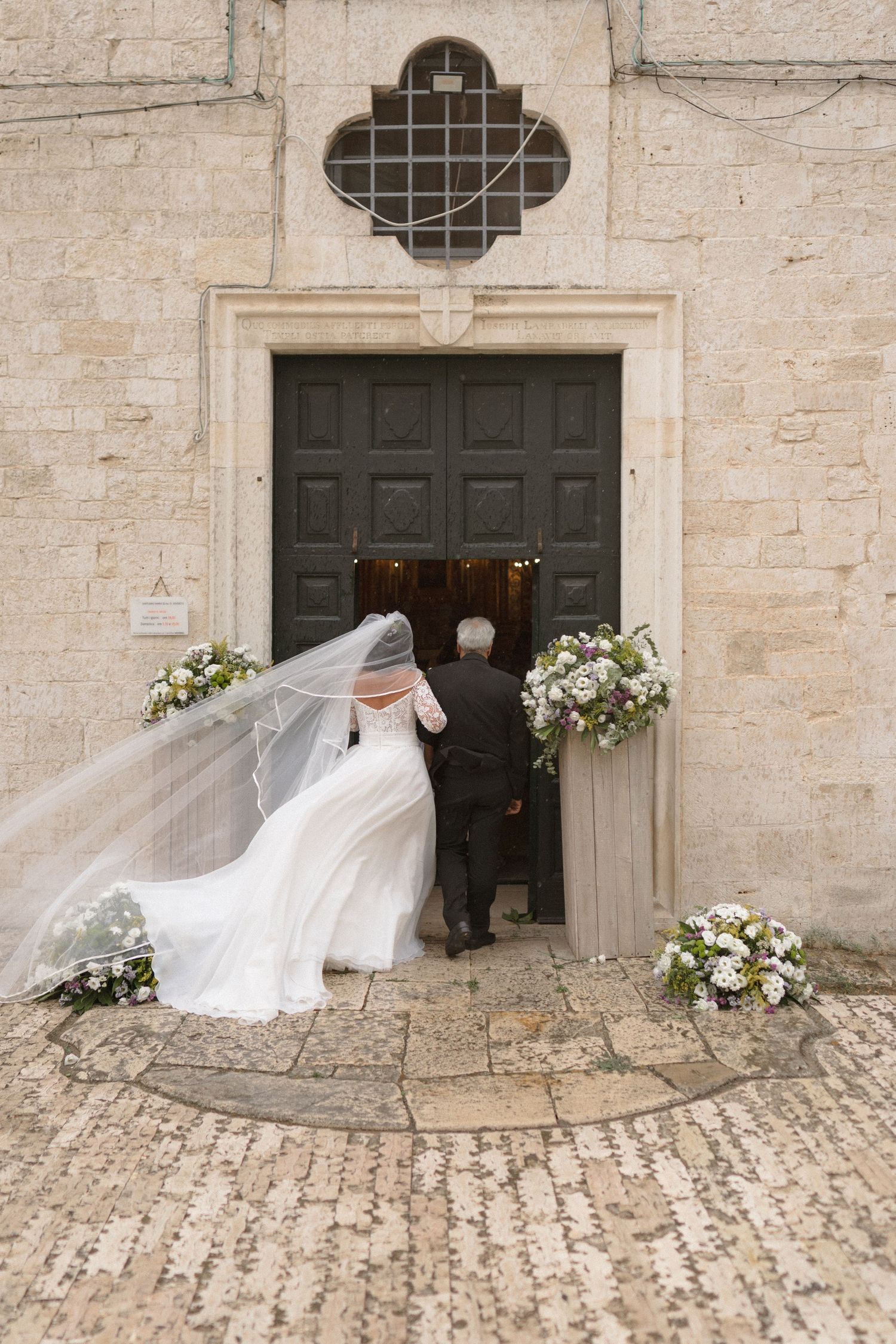 A flowing white wedding veil blows dramatically in the wind against a rustic stone building entrance.