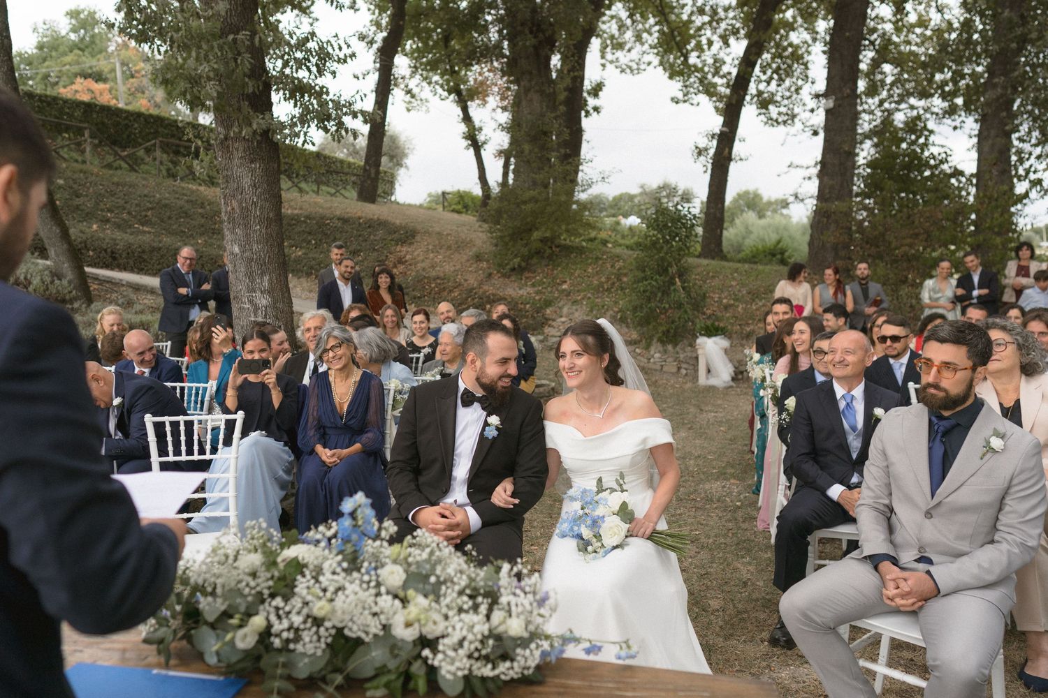 Outdoor wedding ceremony setup with white chairs and floral arrangements in a wooded setting.