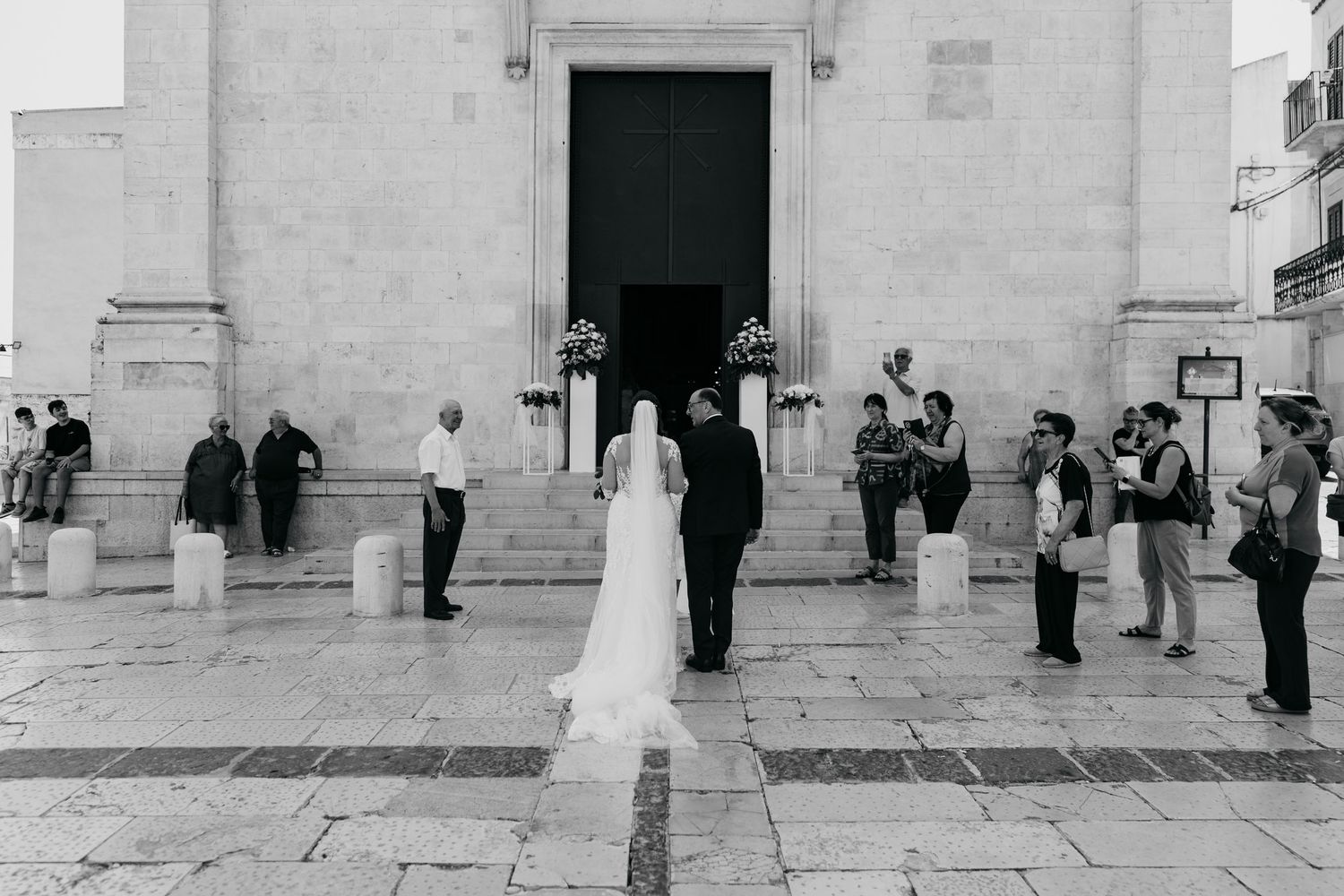 Wedding guests gather outside a white stone church building with large wooden doors.