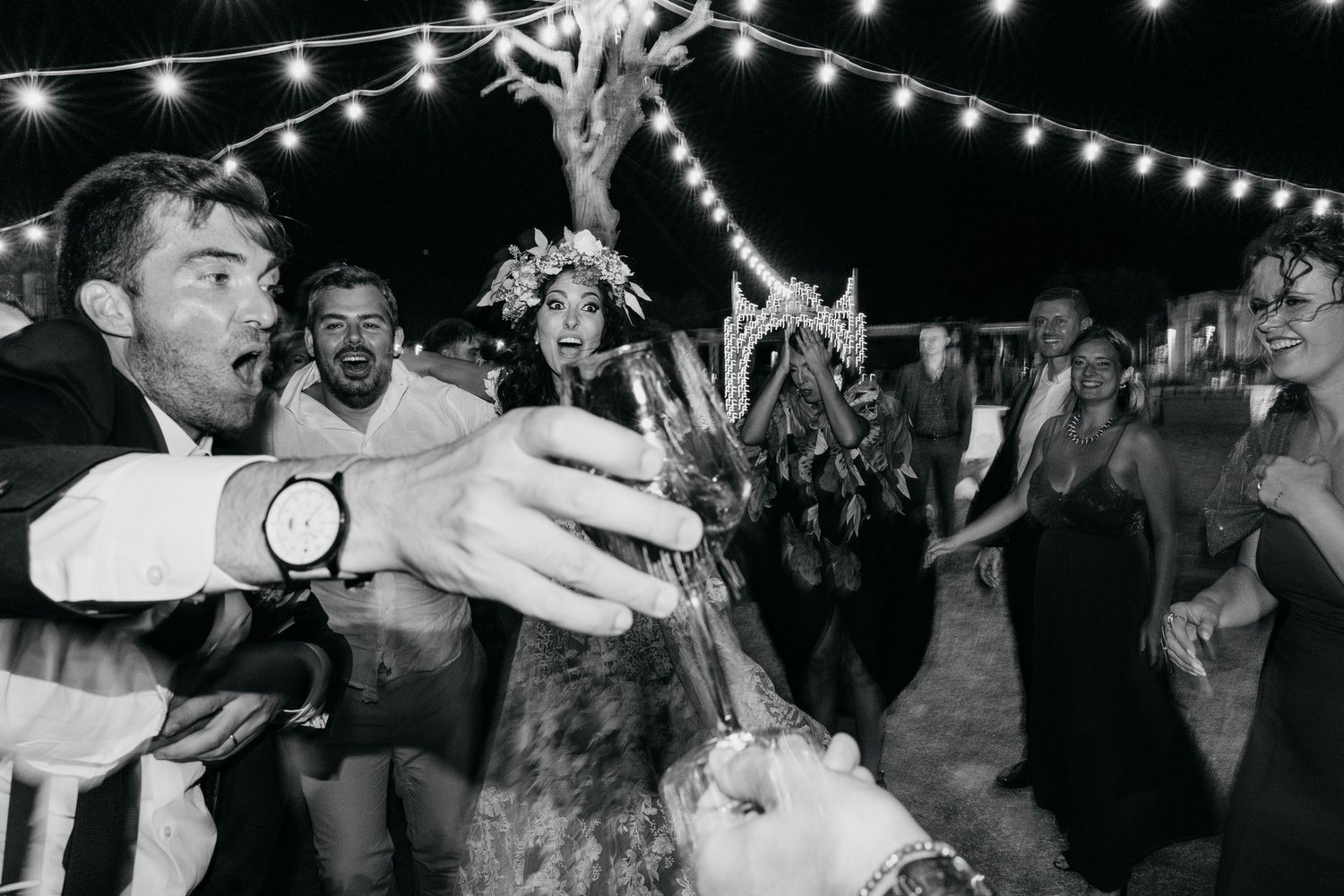 Party guests dancing and celebrating at a lively outdoor wedding reception with string lights overhead.