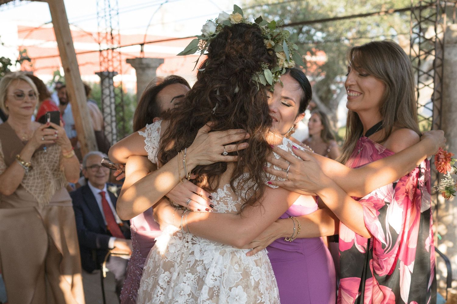 A celebratory group embrace at an outdoor wedding ceremony under a floral arch with natural sunlight.