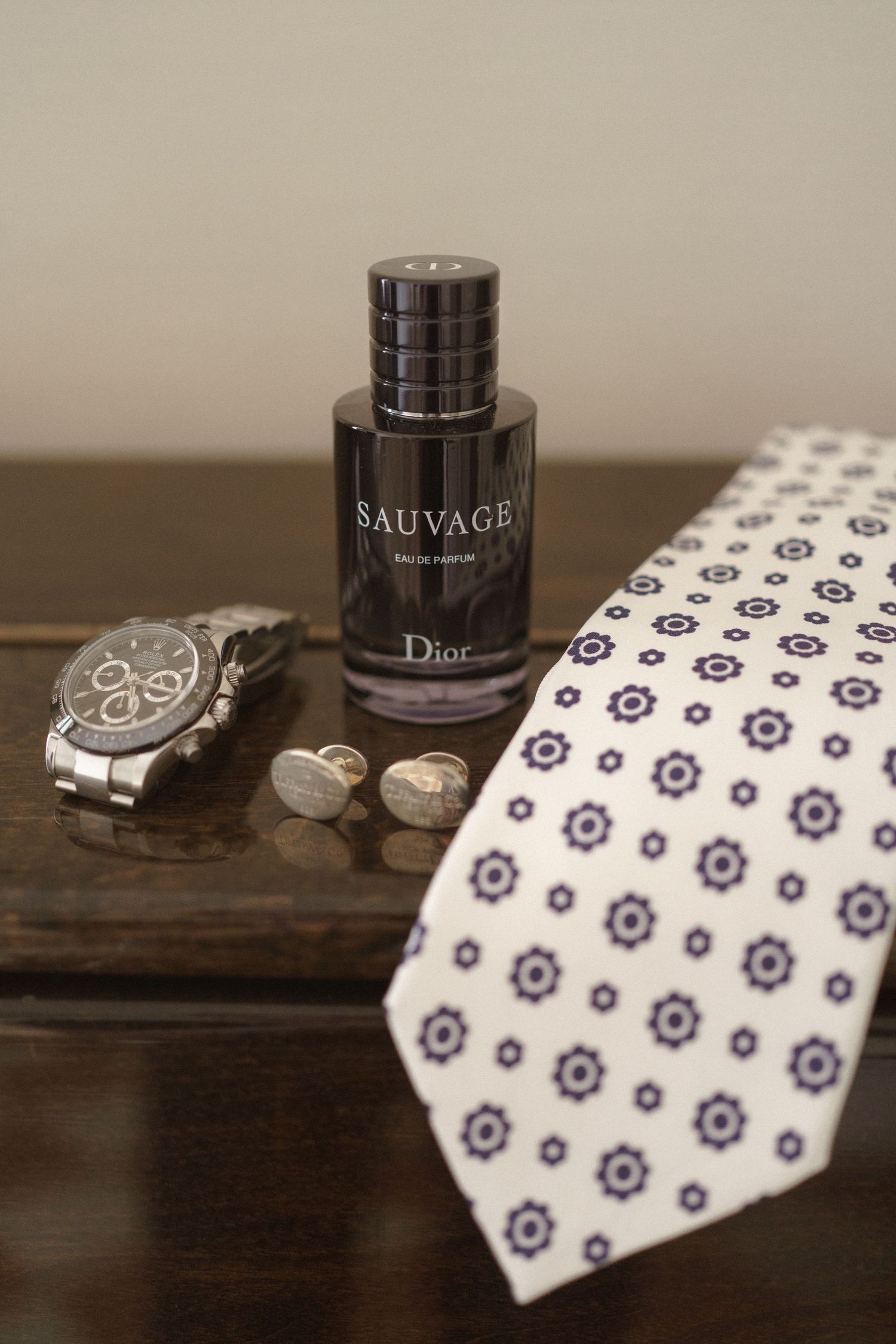 Men's accessories including Dior Sauvage fragrance, silver watch and patterned tie arranged on wooden surface.