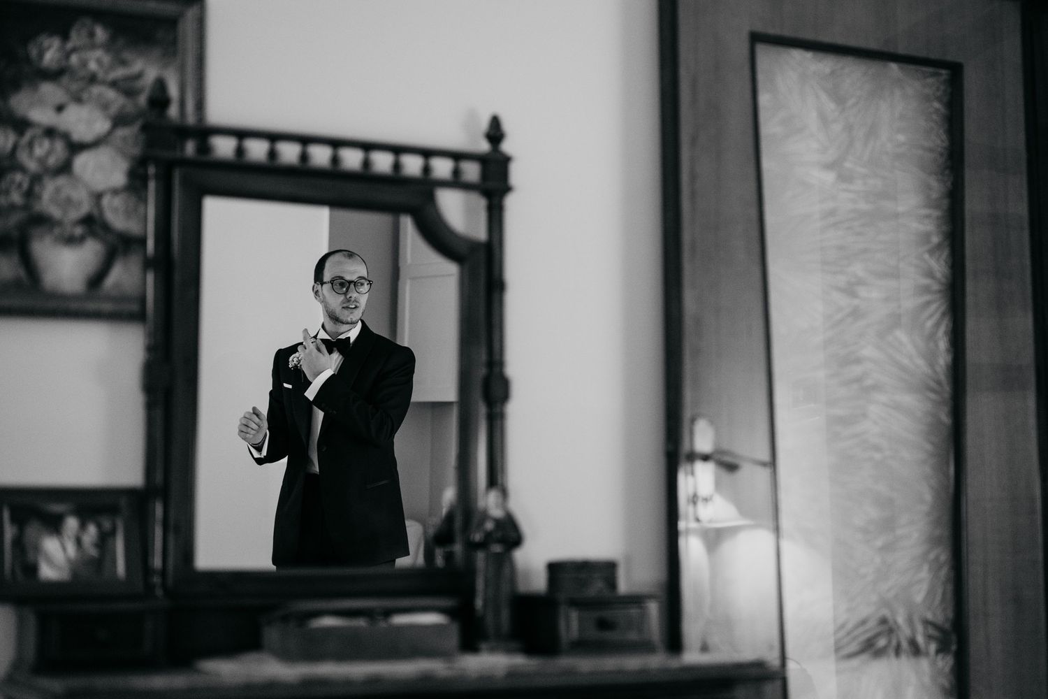 Black and white reflection of a man in formalwear adjusting his bowtie in a bedroom mirror.