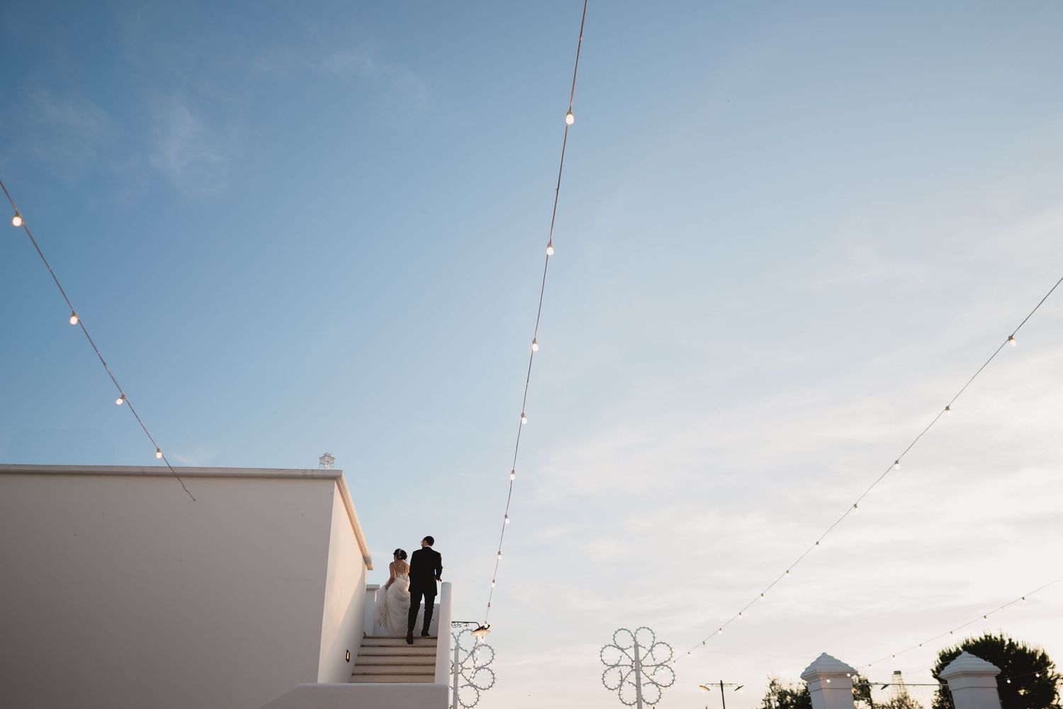 A couple stands on a rooftop with string lights and blue sky in the background.