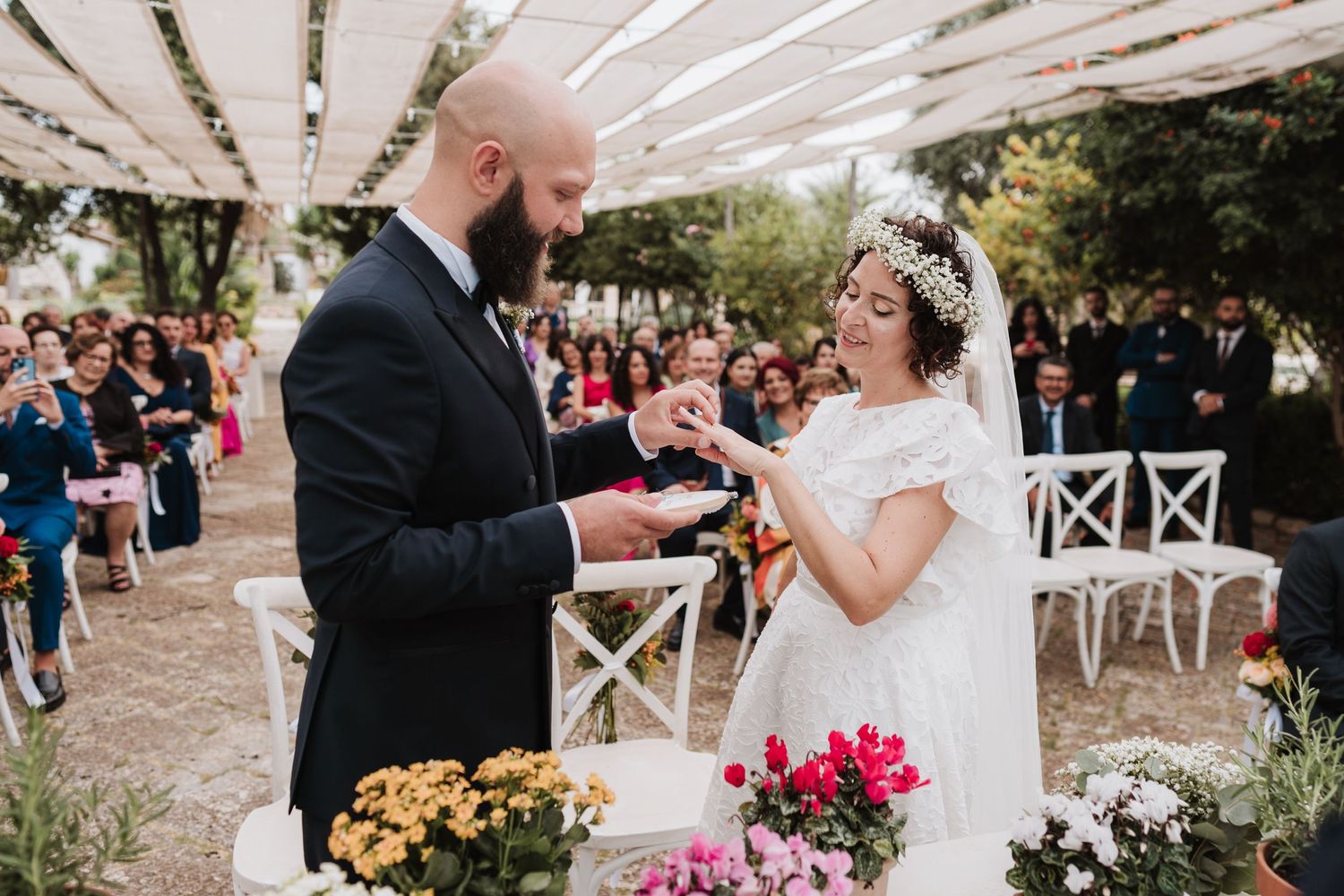 Exchange of rings during outdoor garden wedding ceremony surrounded by colorful floral arrangements.
