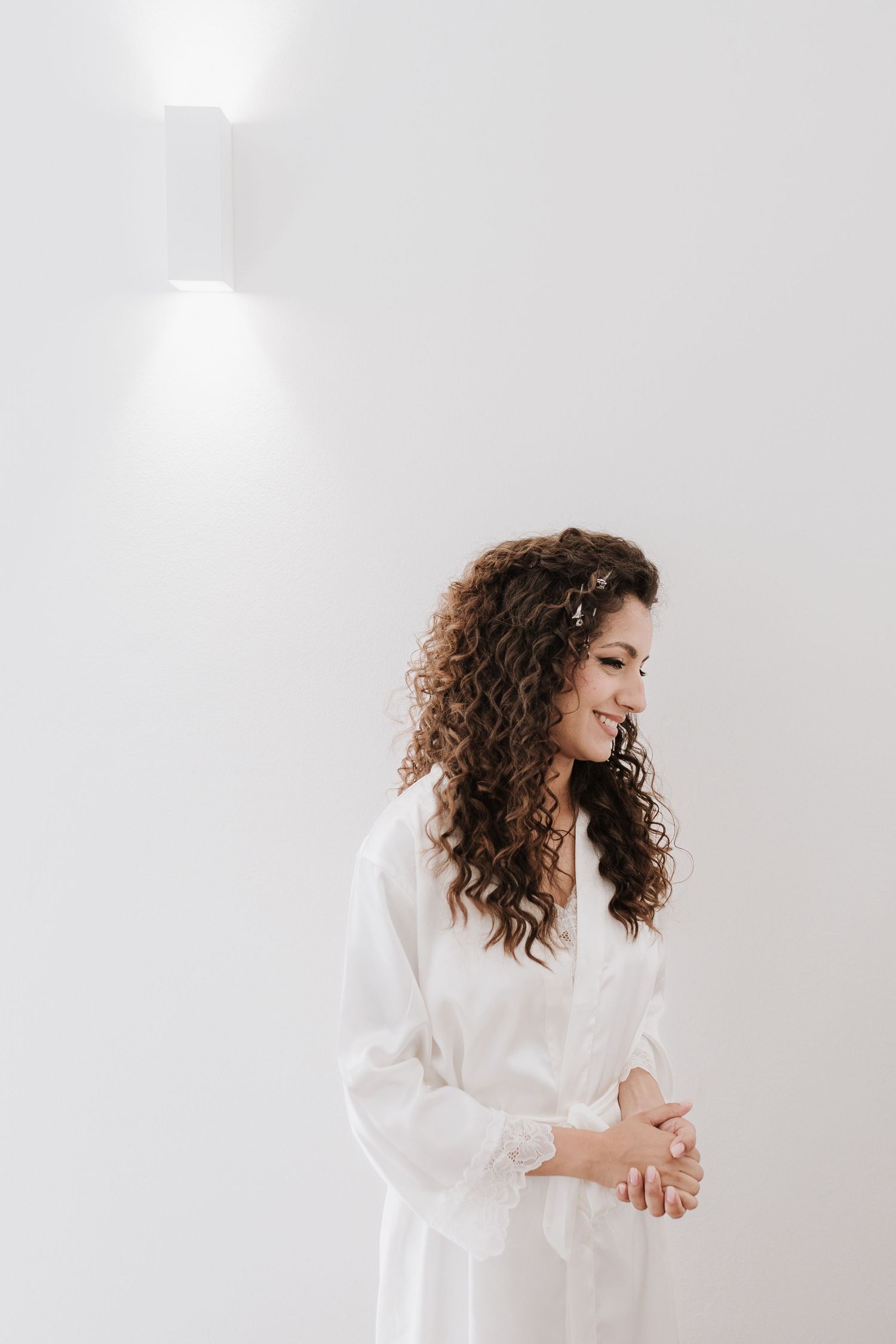 A woman in a white silk robe with long curly brown hair smiles while looking to the side.